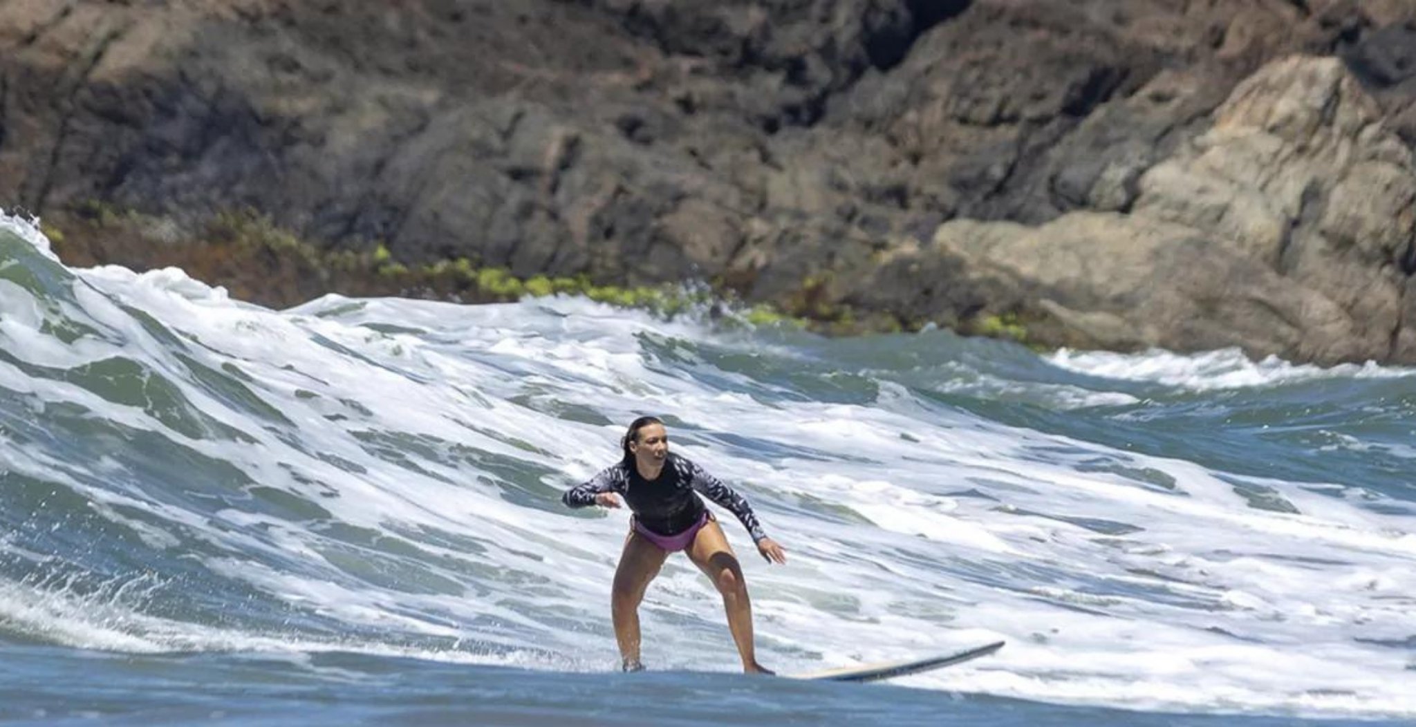 A woman riding a foamy wave on a longboard at a surf school in Itacaré, arms spread wide for balance, with dark volcanic rocks in the background