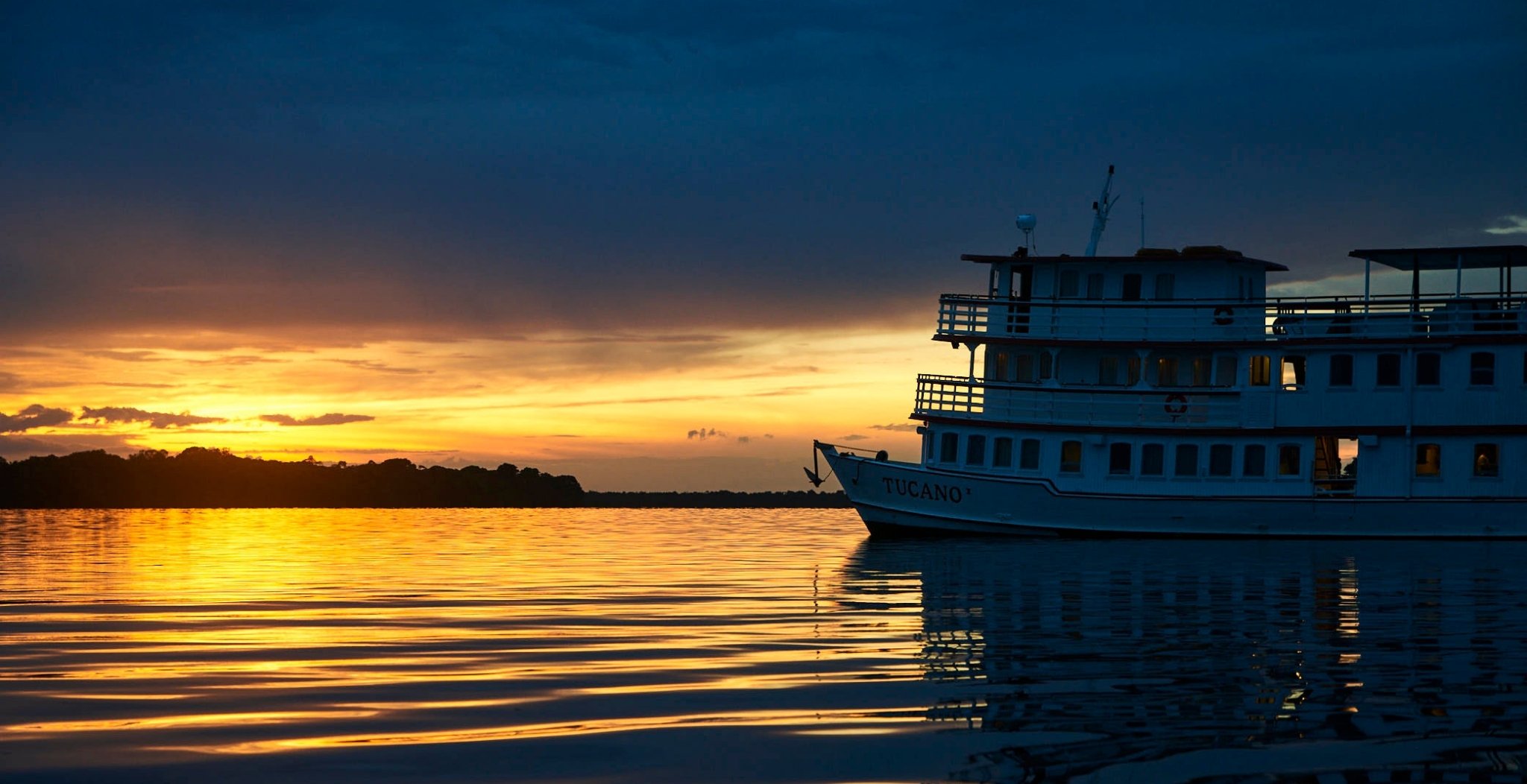 The M/Y Tucano, a white multi-deck river cruise vessel, moored on a calm river at sunset, with a vivid orange and gold sky reflected on the dark water and a treeline silhouetted on the horizon