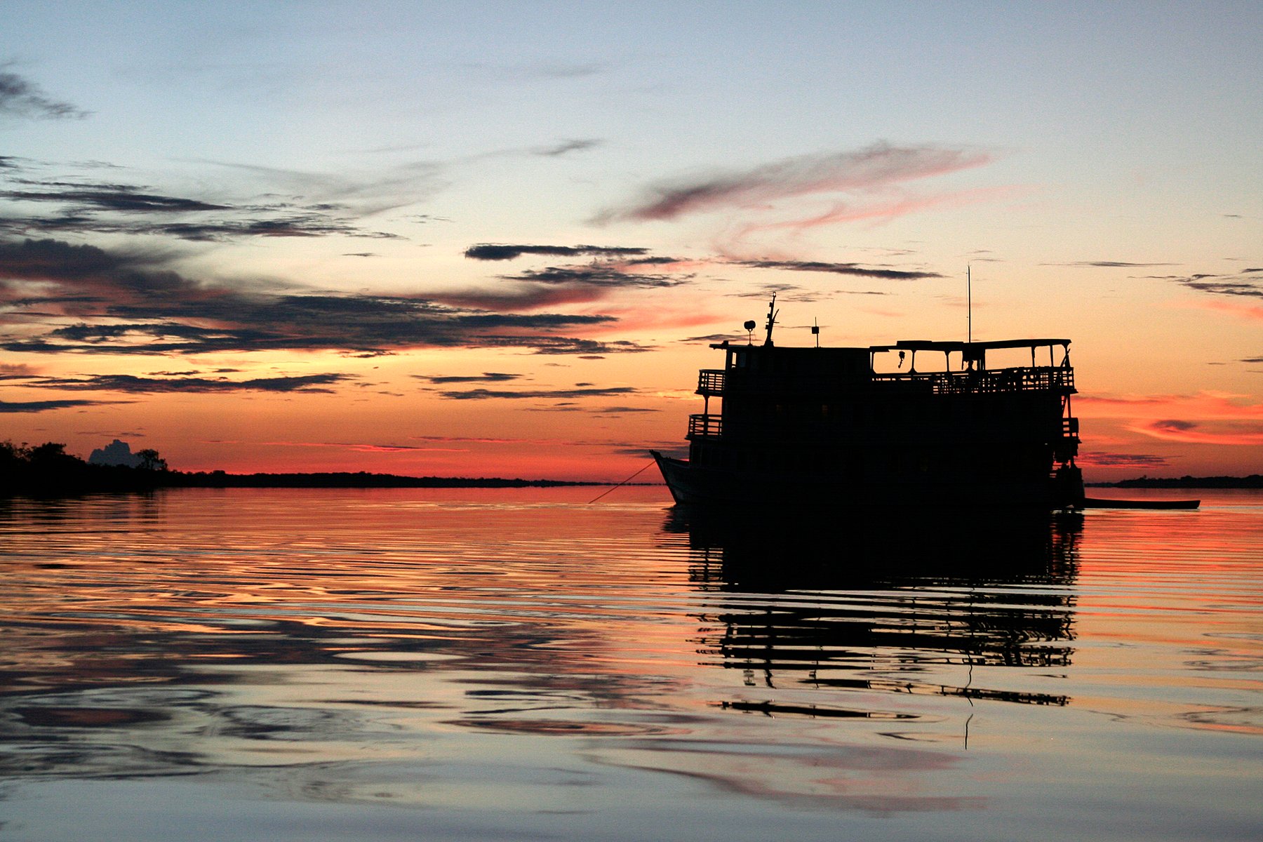 A multi-deck river vessel silhouetted against a vivid orange and red sunset sky reflected on calm dark water, seen during the Tucano Amazon river cruise