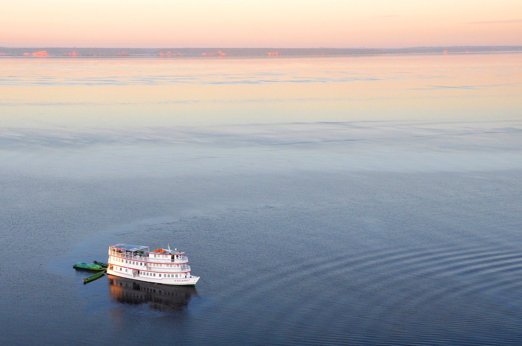 The M/Y Tucano, a small white multi-deck vessel on the Tucano Amazon river cruise, glides across a vast, glassy river at dusk, its wake fanning out behind it beneath a soft pink and peach sky