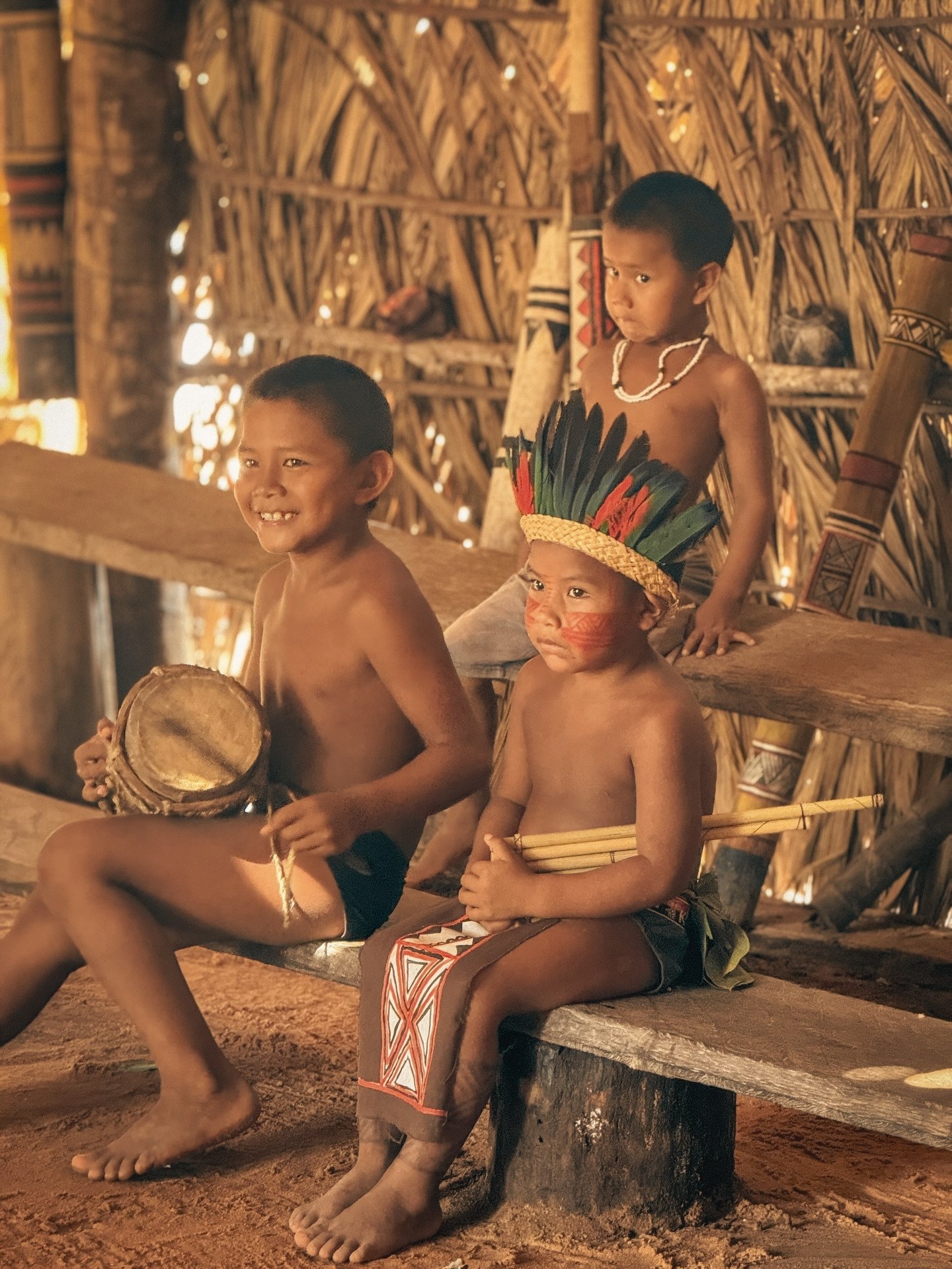 The rhythm starts young out here, and these kids already know every beat. Three Indigenous children inside a thatched-roof maloca on the Ariaú River during an Amazon Cruise Trip in Brazil — one boy smiles holding a small hand drum, another wears a feathered headdress and red face paint, a third watches quietly from behind