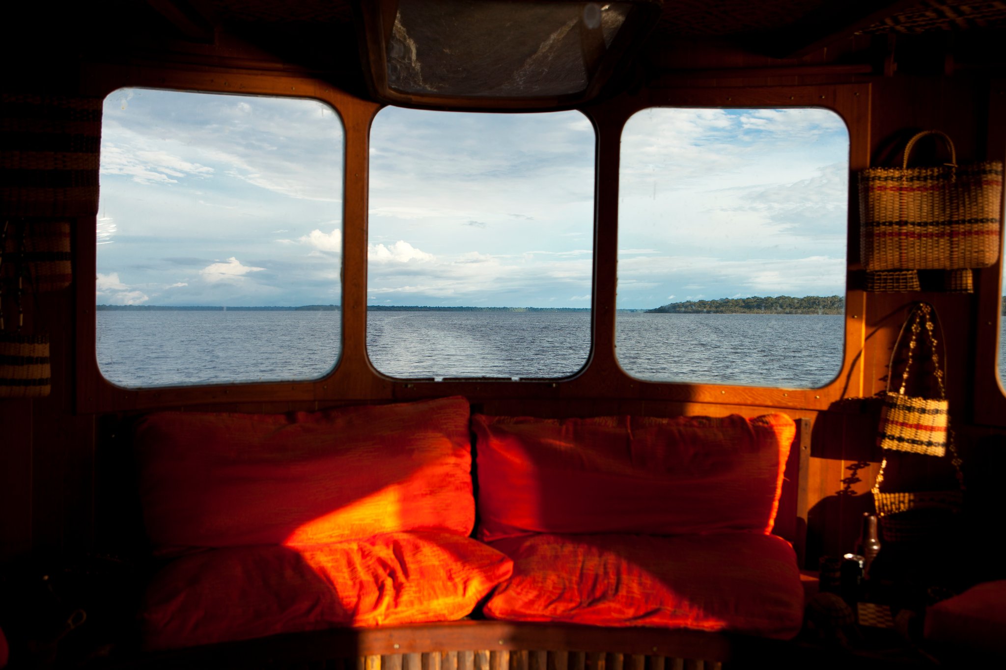Your living room for the next few days happens to have a very good view. Warm red cushions on a wooden settee inside an Amazon jungle cruise Brazil riverboat salon, with three panoramic windows framing a wide river and forested horizon beyond