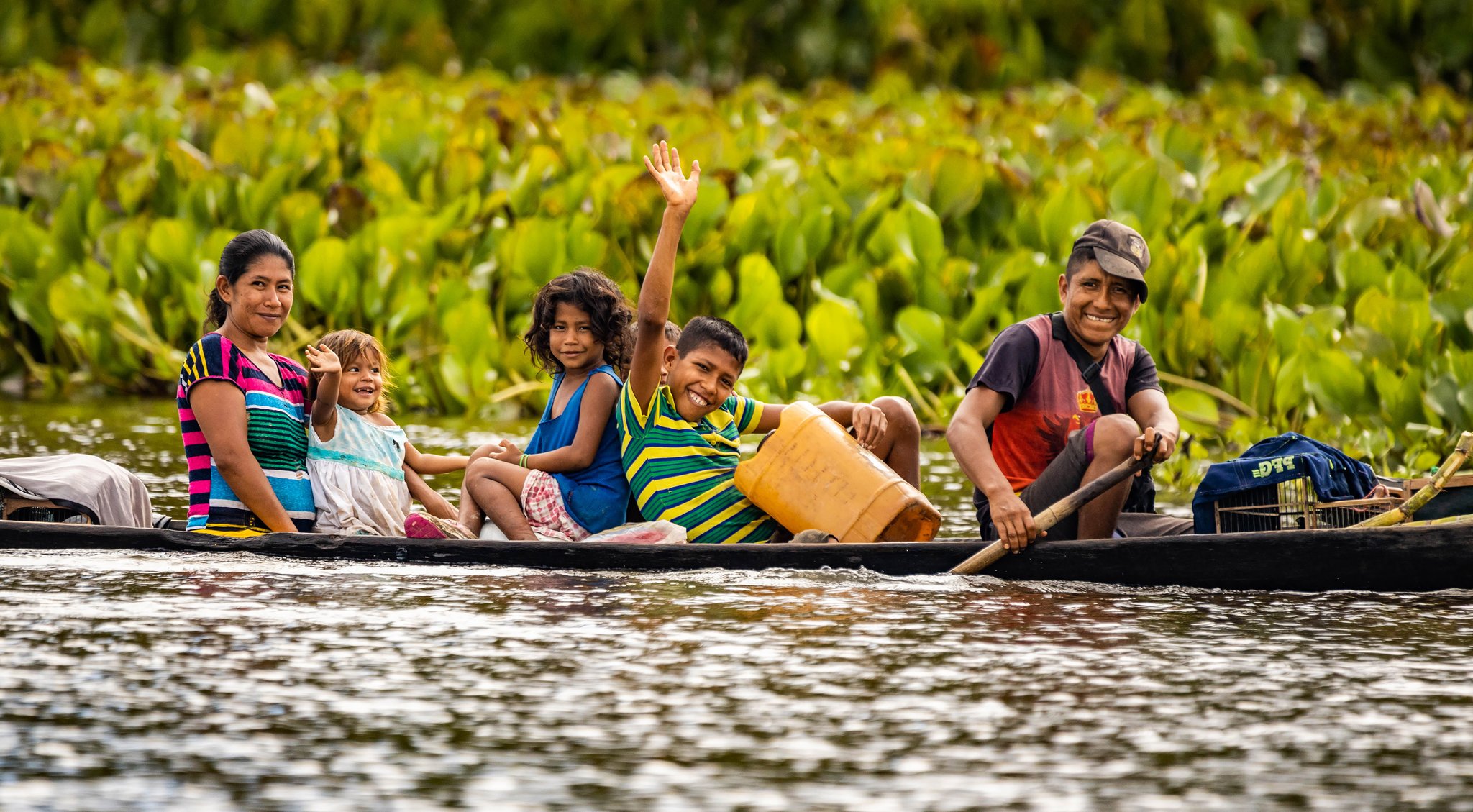 Vijf mensen, één kano, en een groet die je bijblijft. Een glimlachende lokale familie — moeder, vader en drie kinderen — peddelt in een houten kano langs weelderige groene waterplanten op een rustige Amazone-rivier, terwijl een jongen vrolijk naar de camera zwaait