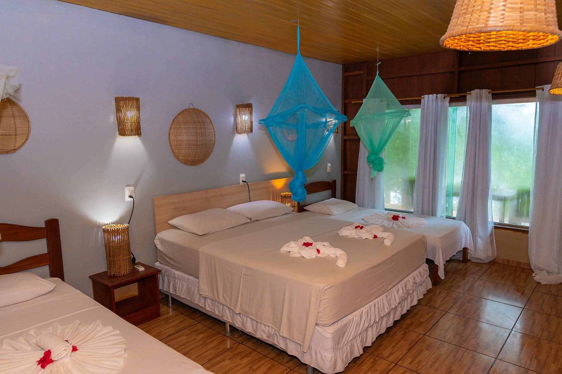 Handdoekzwanen, klamboes en het oerwoud op een steenworp afstand. Een driepersoonsslaper in de Amazon Turtle Lodge met kleurrijke hangende klamboes, gevouwen handdoekdecoraties op witte beddengoed, gevlochten rotanlampen en een raam met uitzicht op weelderig groen