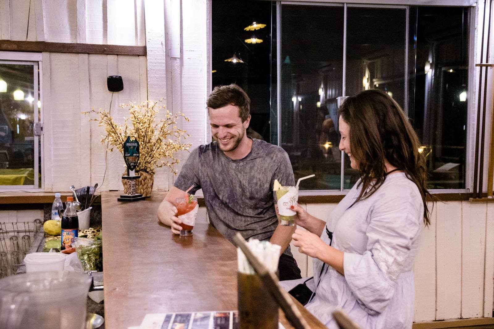 Twee reizigers lachen en houden cocktails vast aan een rustieke binnenbartoonbank tijdens een budget Pantanal-tour, met warm licht en een Pantanal biertap op de achtergrond