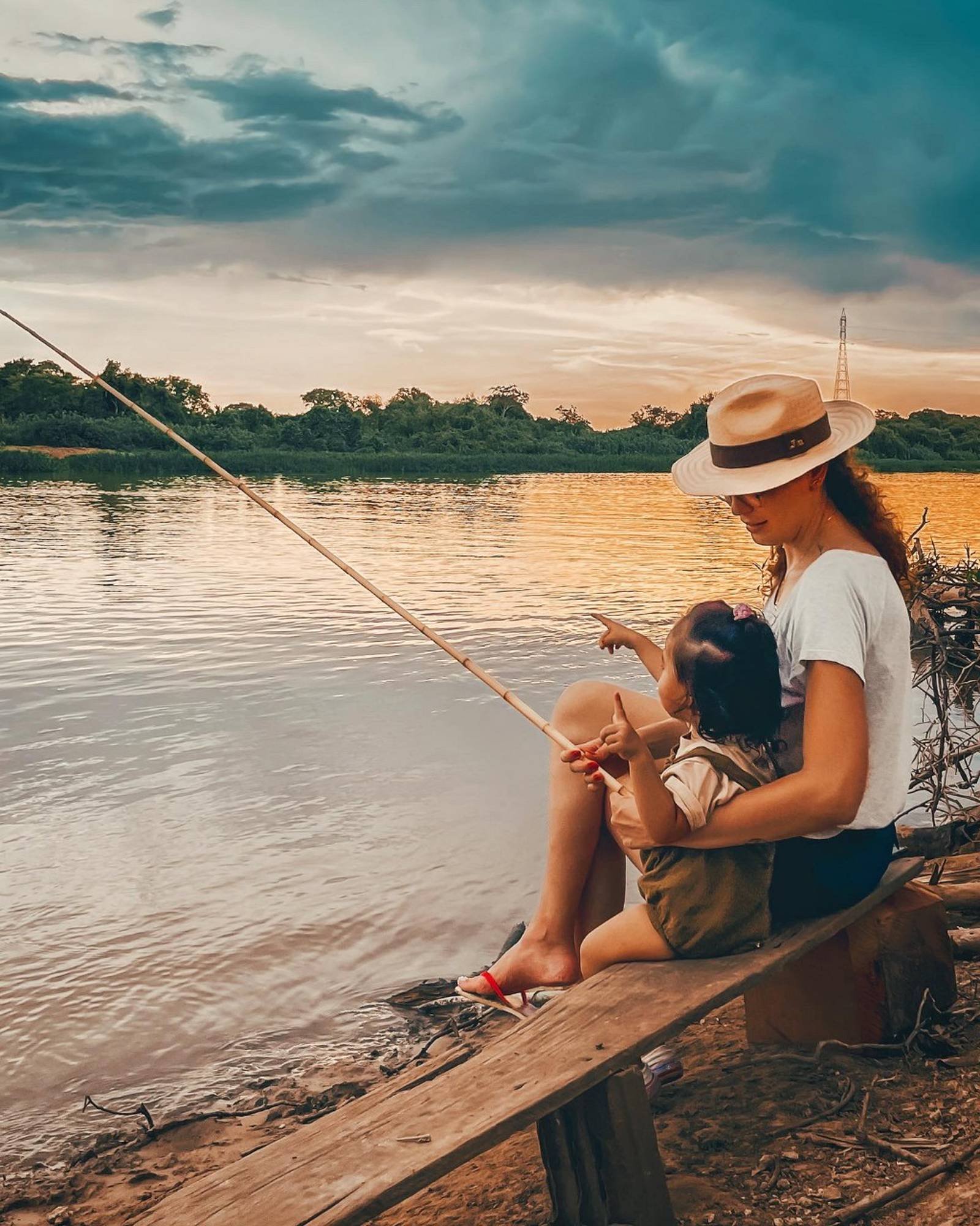Een vrouw met een panama hoed houdt een peuter op schoot en vist met een bamboehengel aan de rivieroever bij zonsondergang, met gouden licht dat weerkaatst op het stille water