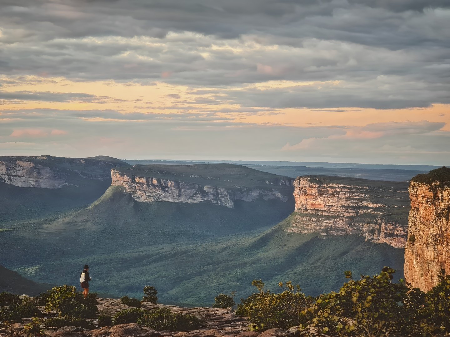 Een eenzame wandelaar staat op een rotsrichel met uitzicht op de uitgestrekte zandstenen tafelbergen en groene valleien tijdens een Chapada Diamantina trip in het schemer, met warm licht dat door dramatische wolken breekt