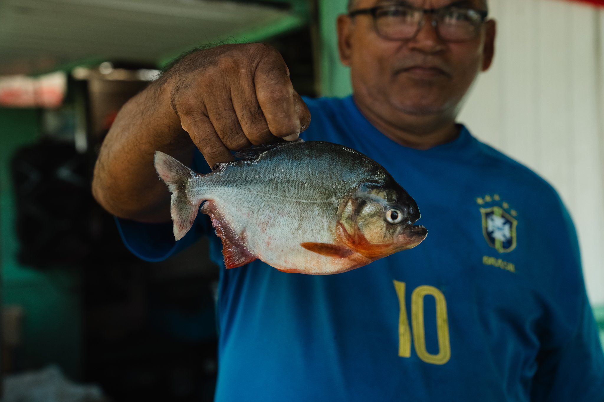 A man in a Brazil football shirt holds up a freshly caught piranha with vivid red-orange fins, a direct example of community based tourism where local knowledge and river life are shared with visitors