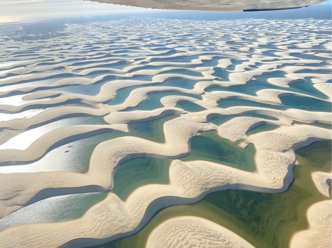Aerial view from a small aircraft over the Lençóis Maranhenses full itinerary landscape, showing vast white sand dunes interlaced with turquoise and emerald seasonal lagoons stretching to the horizon