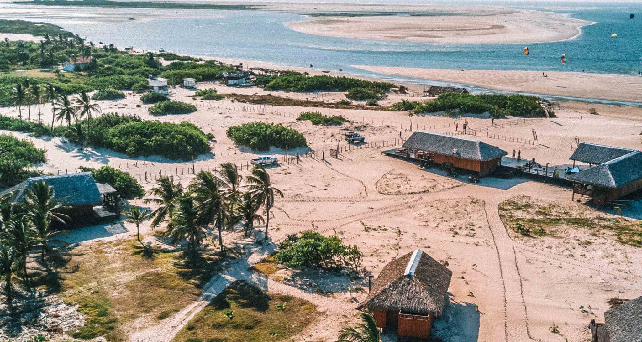 Aerial view of thatched-roof huts, palm trees, and sandy tracks in the rustic village of Atins, with the Lençóis Maranhenses Full Itinerary taking travelers along the coastline where kite surfers dot the blue waters beyond