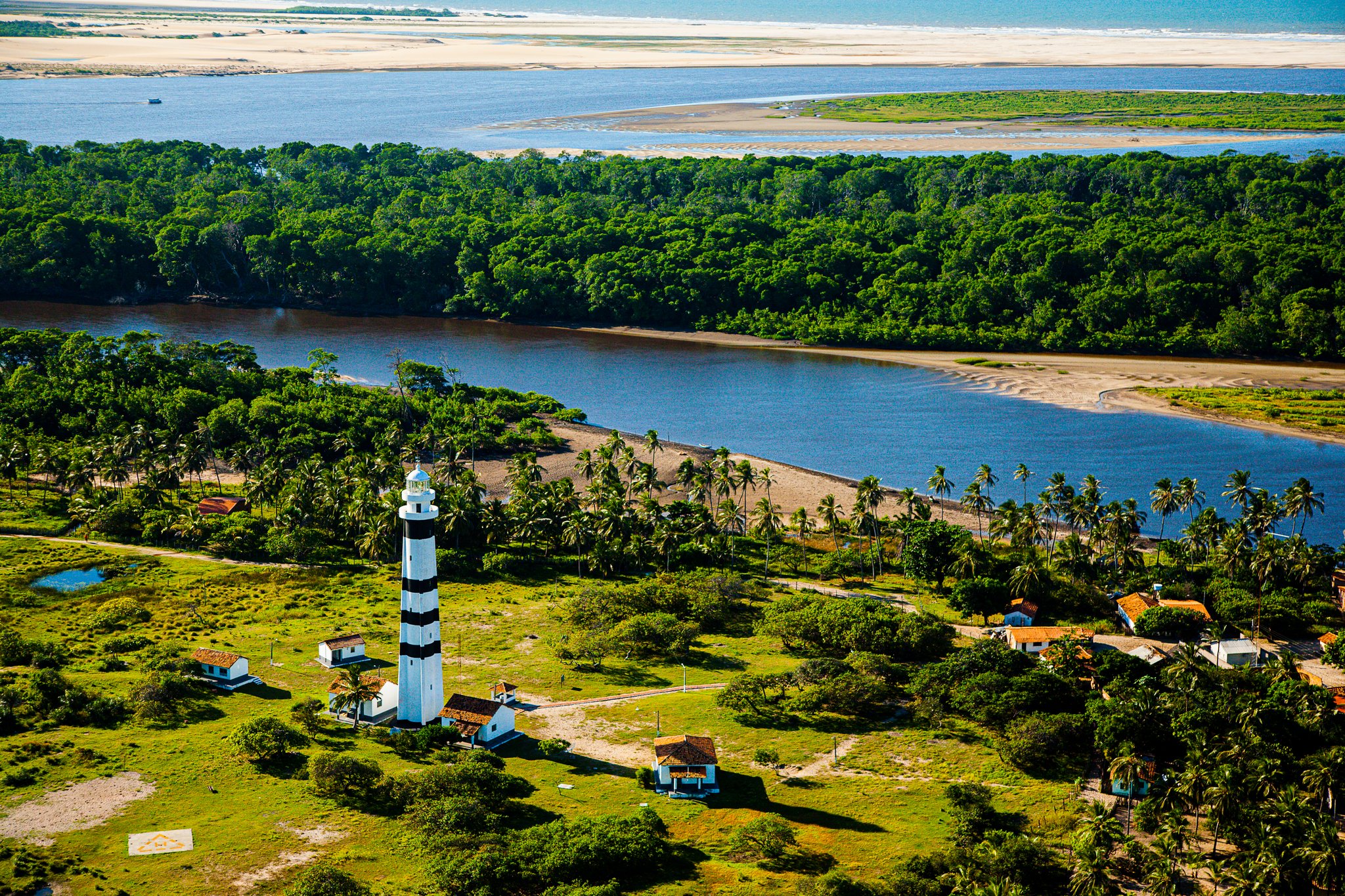 Aerial view of a black-and-white striped lighthouse surrounded by palm trees and small buildings, with the Preguiças River, dense forest, and white sand dunes stretching to the horizon — a landmark stop on the Lençóis Maranhenses Full Itinerary
