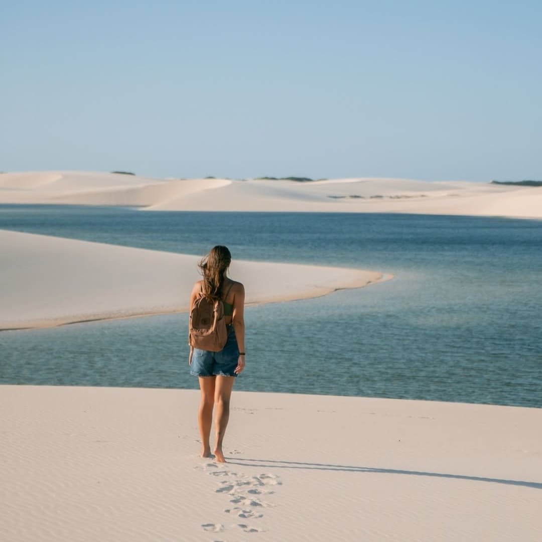 A woman with a backpack walks barefoot across white sand dunes toward a crystal-clear blue lagoon on a Lençóis Maranhenses full itinerary, her footprints trailing behind her
