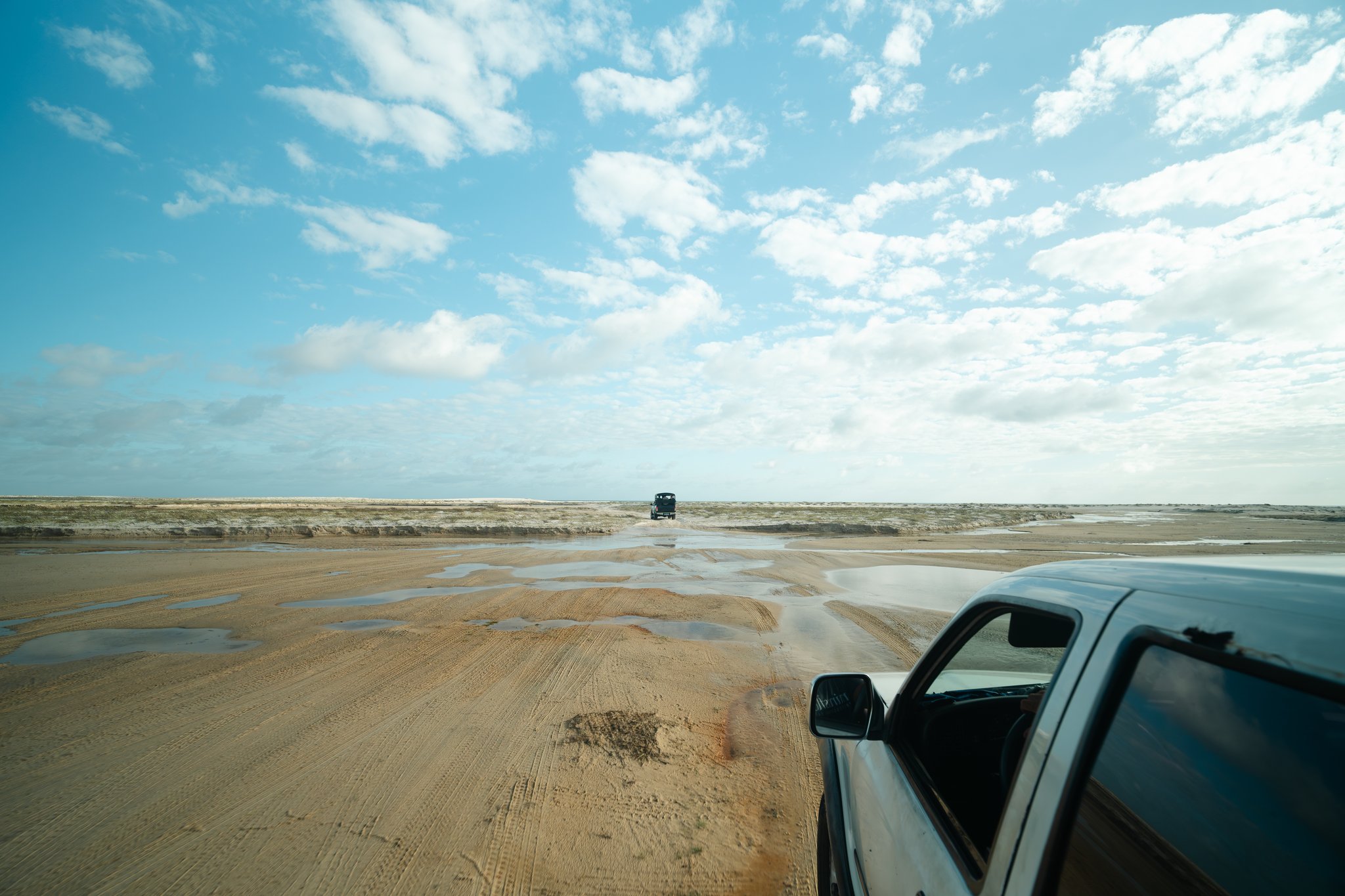 Die Strecke entscheidet das Wasser, nicht die Karte. Zwei Geländewagen überqueren eine weitläufige, nasse Sandebene unter einem teilweise bewölkten blauen Himmel auf dem Lençóis Maranhenses Trek ab Atins, mit flachen Wasserpfützen, die das Licht auf dem Untergrund spiegeln