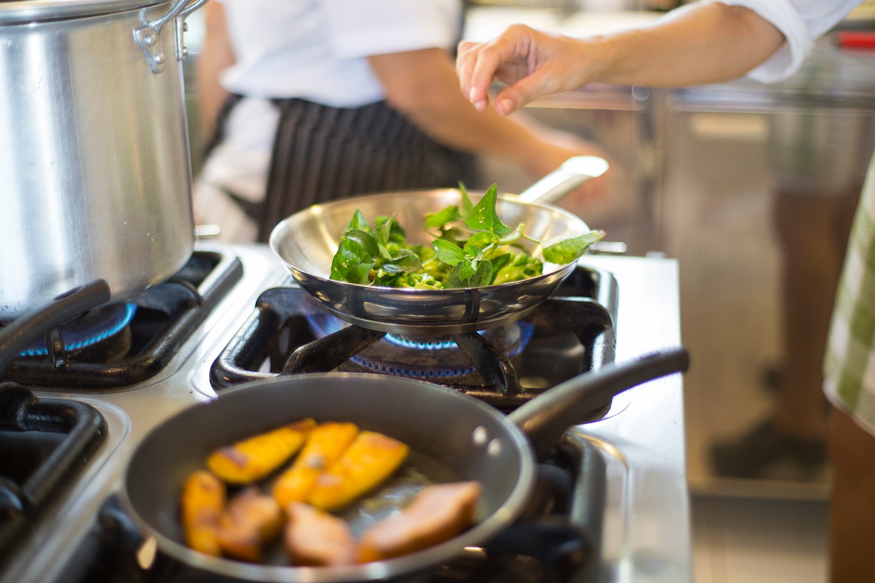 Verse smaken, open vuur en een kok die zijn werk serieus neemt. Een kok in een gestreept schort roerbakt verse groene bladeren in een roestvrijstalen pan op een gaspit in de keuken van Mirante do Gavião lodge, met goudgaar gebakken fruitschijfjes in een tweede pan op de voorgrond