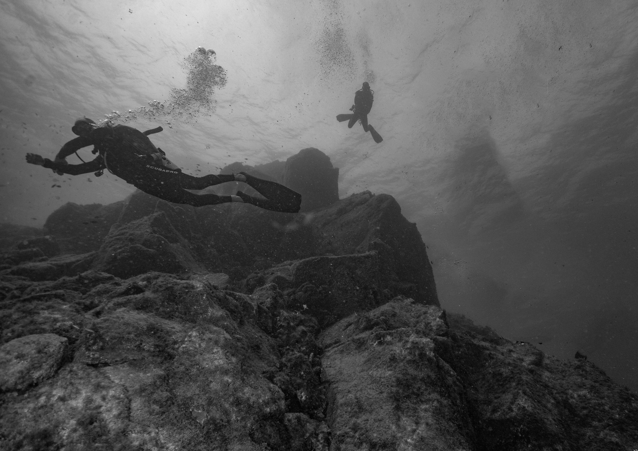 Dois mergulhadores com scuba exploram uma impressionante formação rochosa subaquática durante um curso de mergulho Open Water, com bolhas de ar subindo em direção à superfície, em foto preto e branco tirada de baixo para cima