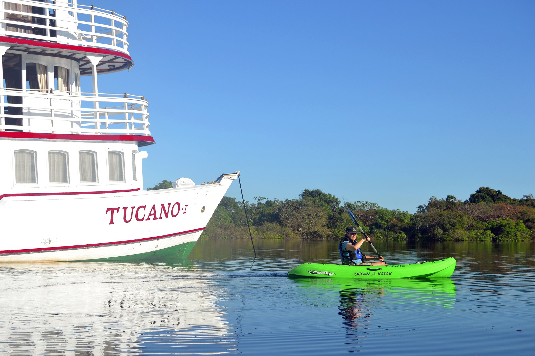 A traveler in a green Ocean Kayak paddles beside the white hull of the M/Y Tucano I on a calm river, with dense rainforest lining the far bank under a clear blue sky, on a tucano amazon river cruise excursion.