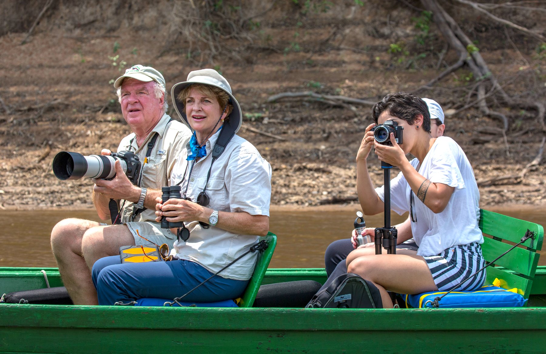 Three travelers on a green wooden boat along a river bank during a Tucano Amazon river cruise, one man holding a telephoto camera lens, a woman with binoculars, and a younger person shooting with a camera on a monopod