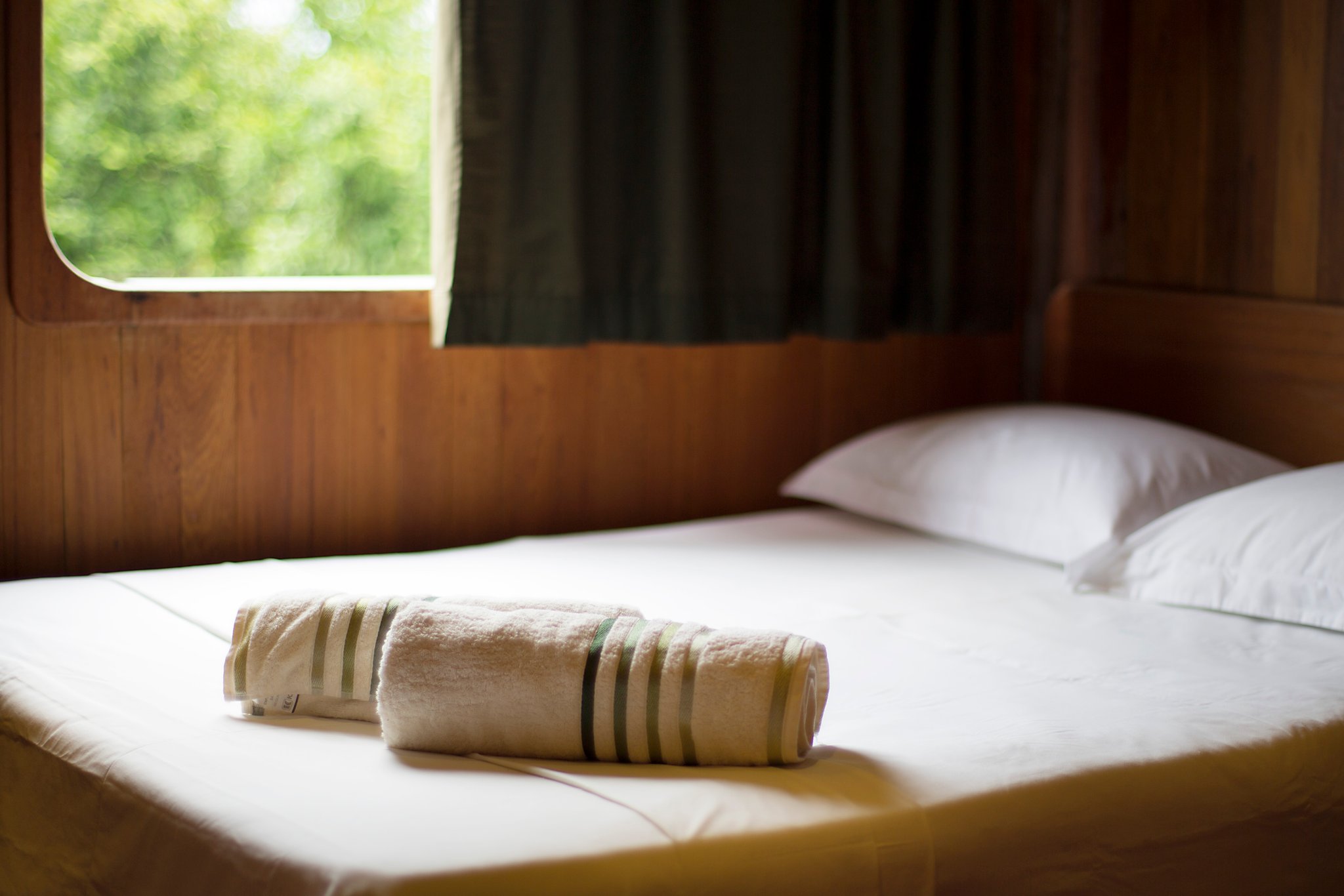 Your floating home for four nights, ready before you even arrive. Neatly rolled striped towels placed on a freshly made bed inside a wood-panelled cabin on an Amazon jungle cruise Brazil, with lush green foliage visible through the porthole window