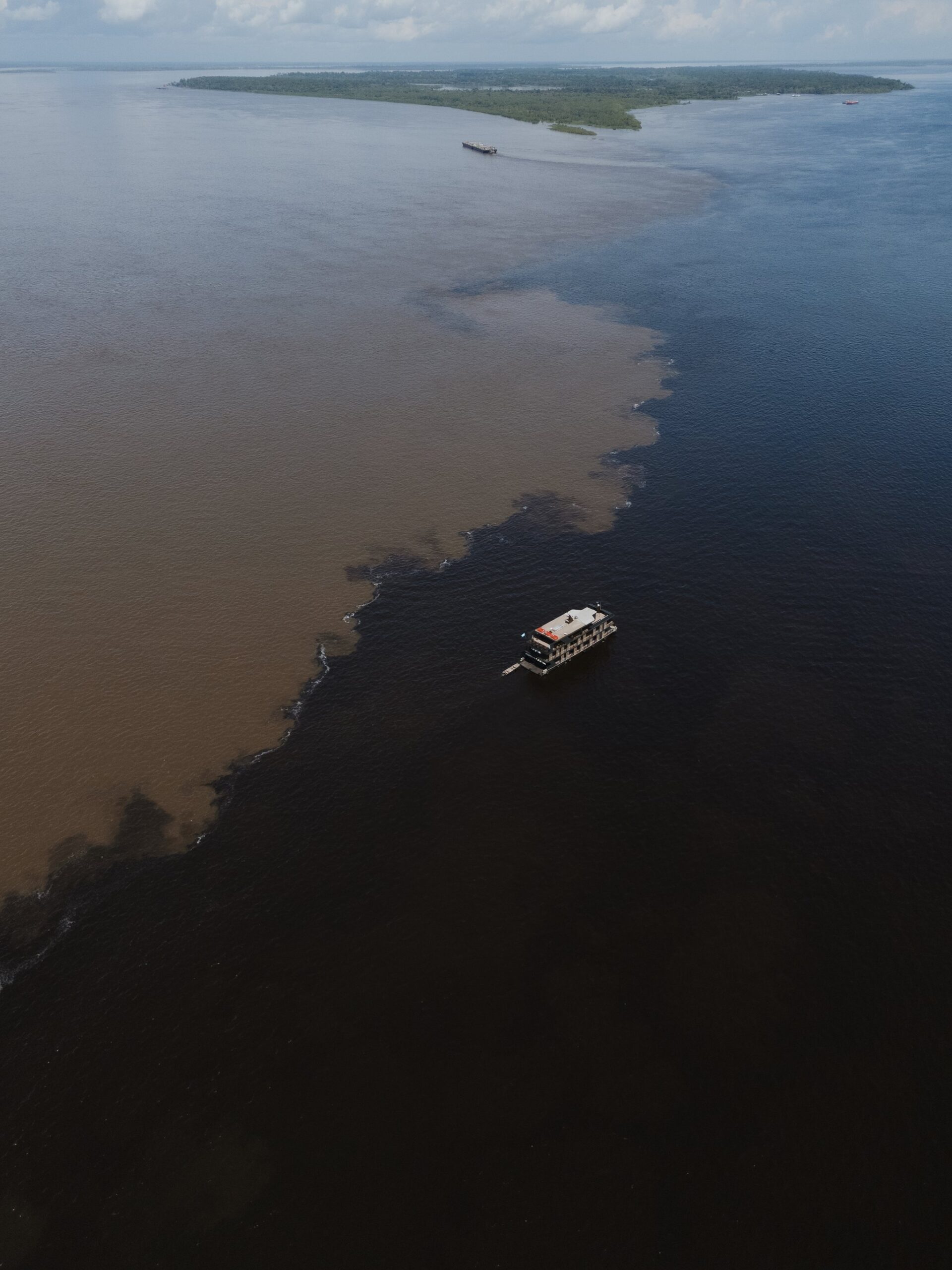 Zwart links, bruin rechts, de natuur trekt hier haar eigen grens. Luchtfoto van een rivierschip dat de Ontmoeting der Wateren oversteekt, waar zwart en bruin water in een grillige grens samenkomen, met een bebost eiland op de achtergrond
