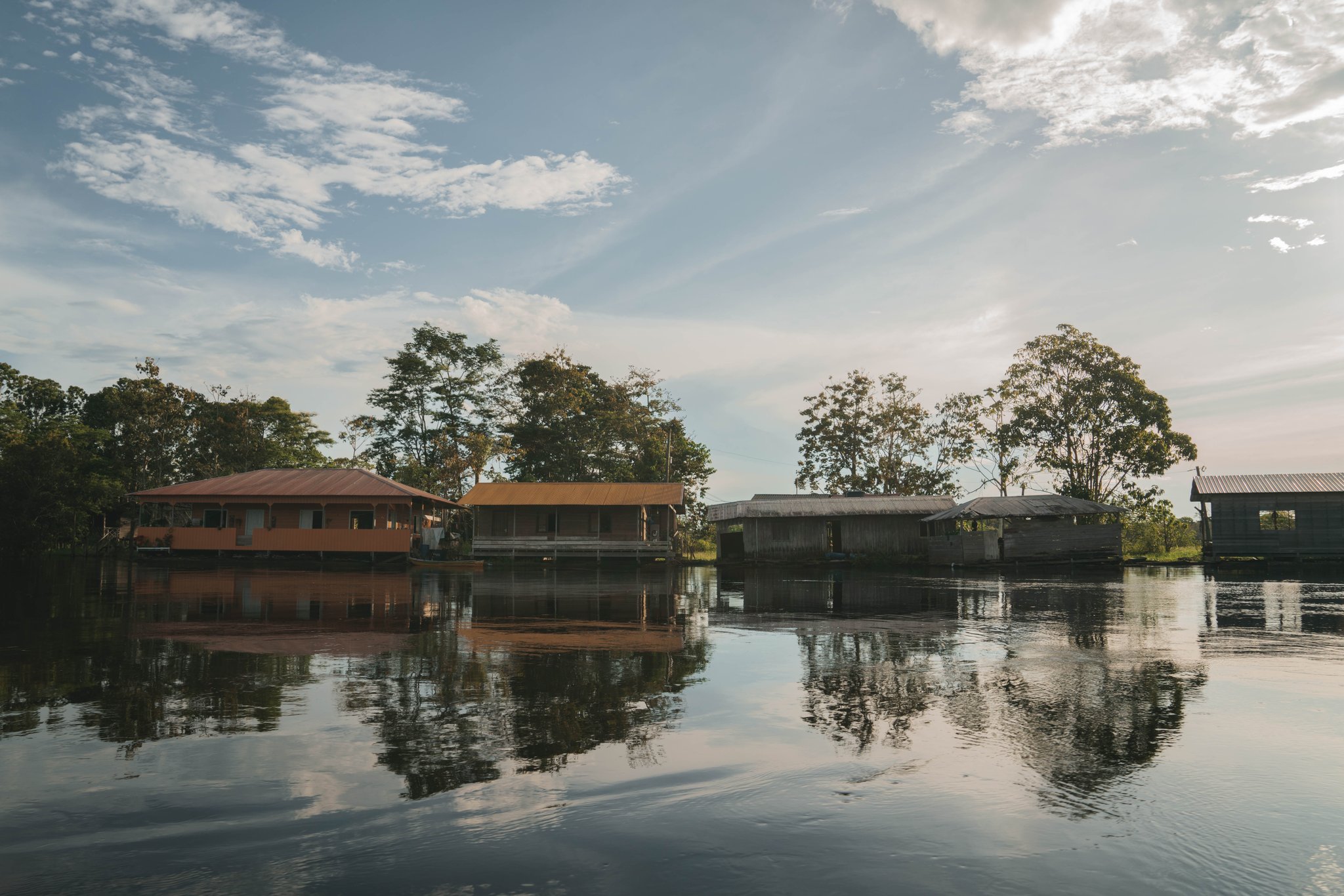 Rust op het water, met het regenwoud vlak achter de veranda. Houten en geschilderde gebouwen op palen van de Amazon Turtle Lodge die direct boven een kalme, donkere rivier staan, met hoge regenwoudbomen weerspiegeld in het stille water onder een gedeeltelijk bewolkte hemel