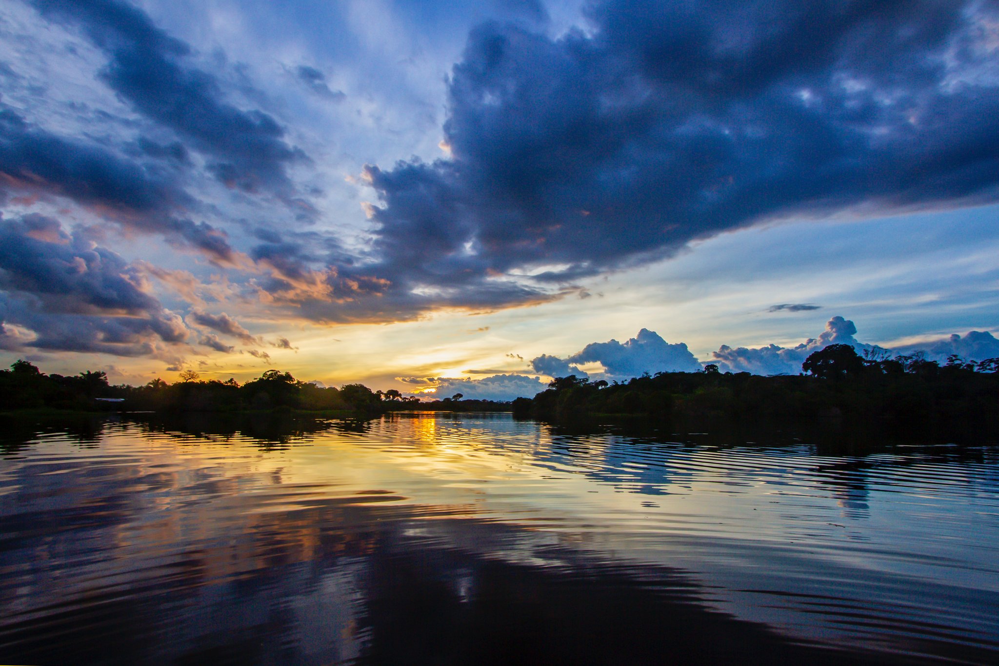 Hier besef je hoe groot de lucht eigenlijk is. Dramatische zonsondergang boven een breed regenwoudrivier, goudkleurig licht weerspiegeld op zachtjes rimpelend water, donkere boomsilhouetten aan de horizon