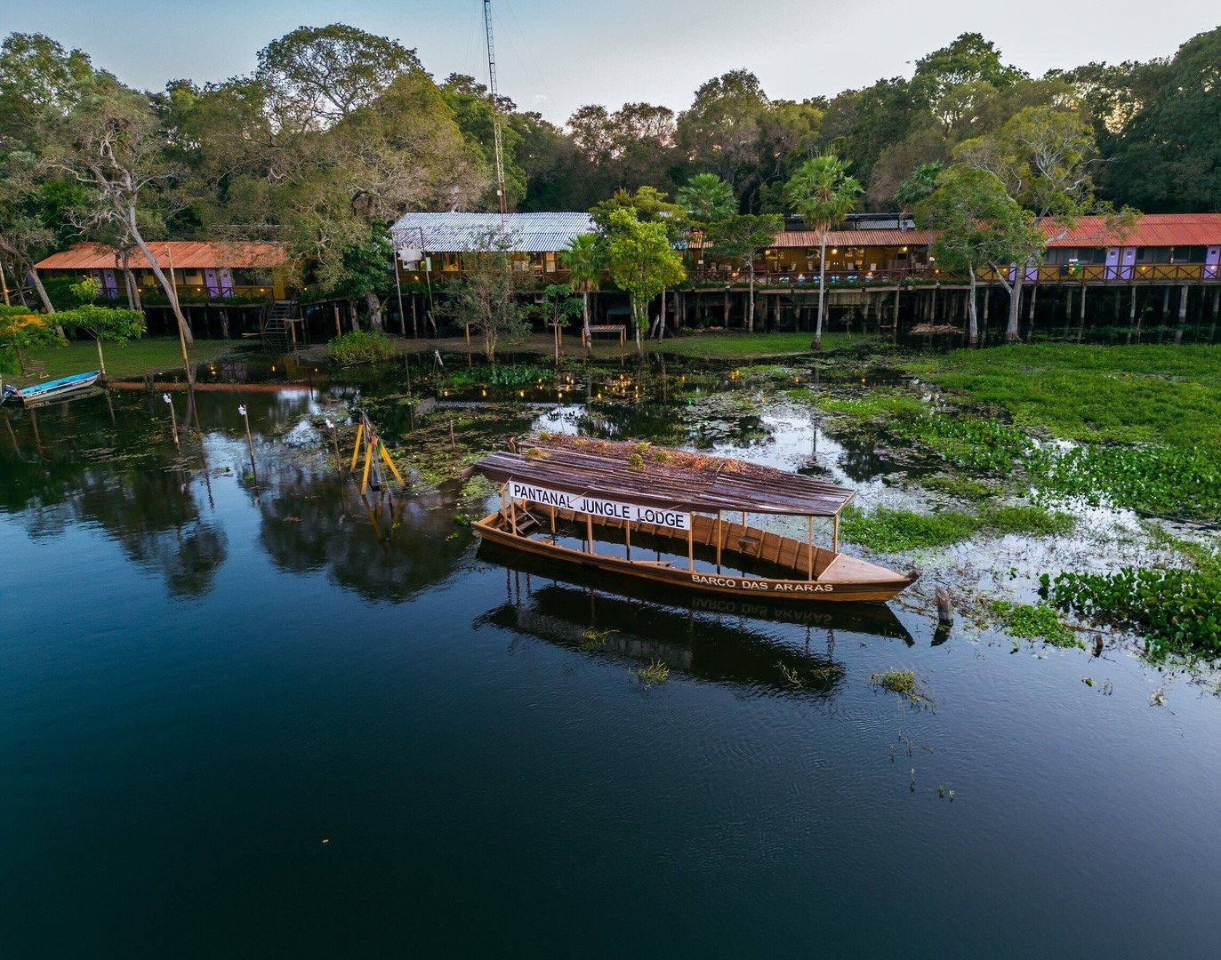 Luchtfoto van het Pantanal Jungle Lodge aan een rustige waterweg, met de houten rondvaartboot 'Barco das Araras' afgemeerd tussen waterplanten en paalwoningen verscholen in dicht groen