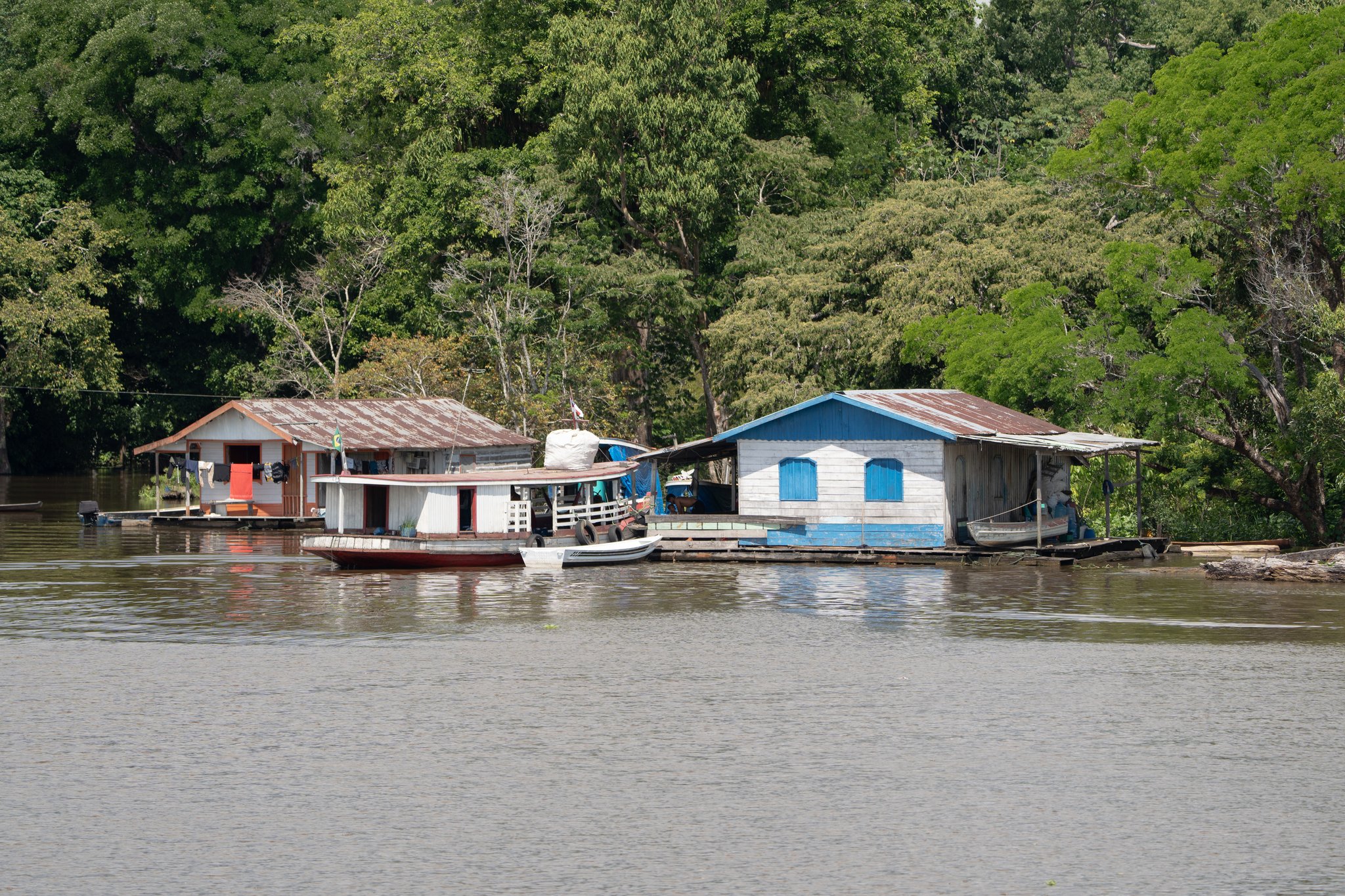 Wooden riverside homes with corrugated tin roofs and small boats moored at a floating dock, set against dense tropical forest — a glimpse of community based tourism along an Amazonian waterway