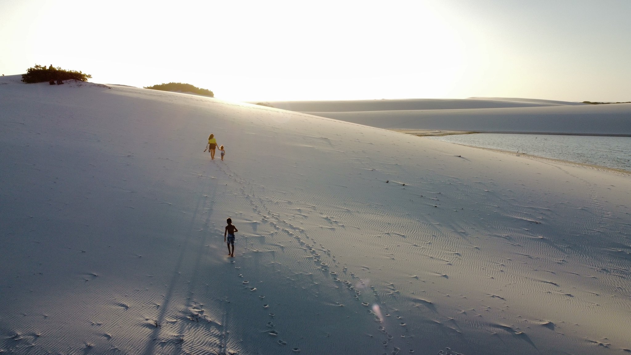 An adult and two children climb a vast white sand dune beside a still lagoon during the golden hour on a Lençóis Maranhenses full itinerary