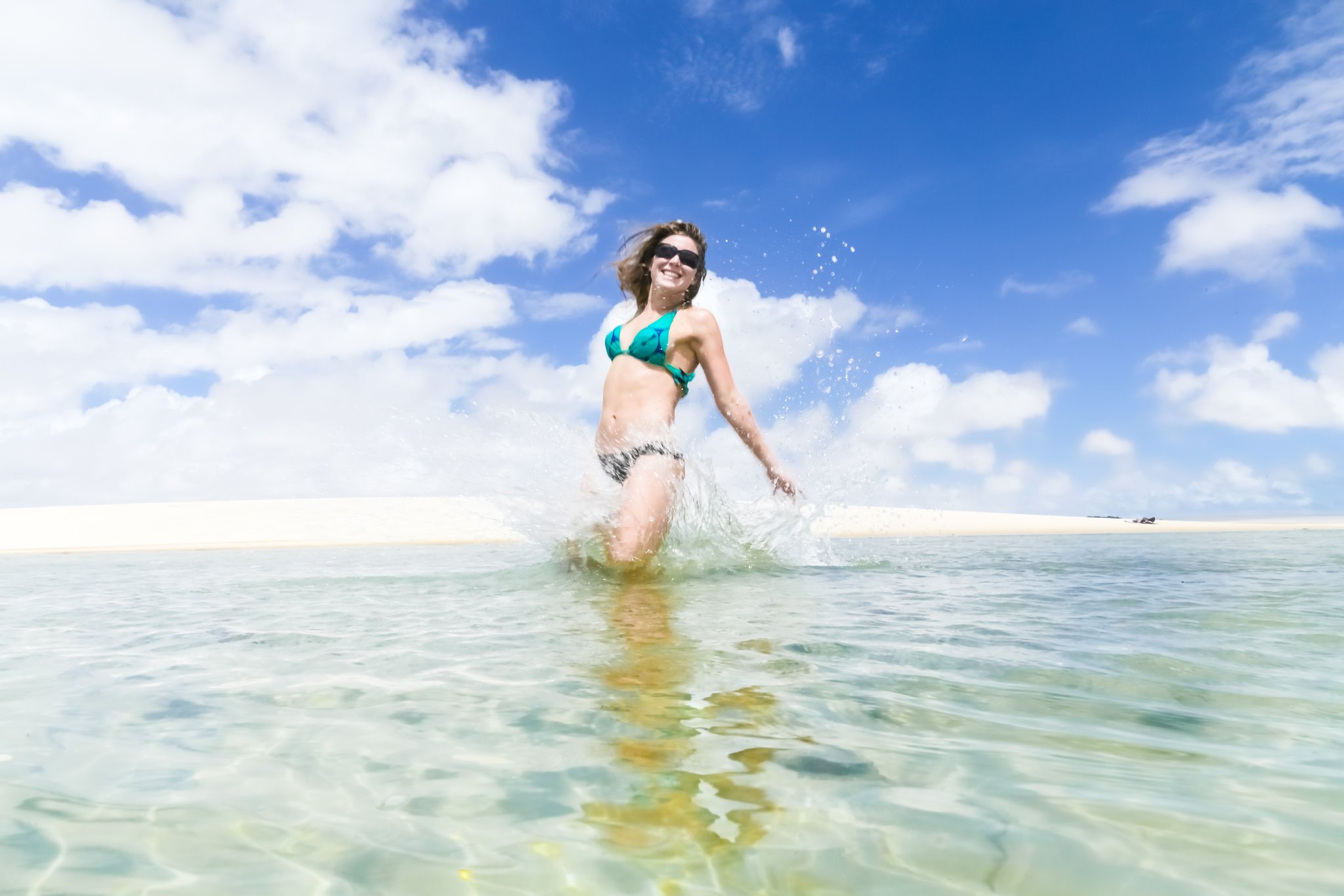 A smiling woman in a teal bikini splashes through crystal-clear shallow lagoon water against white sand dunes and a vivid blue sky in Lençóis Maranhenses Full Itinerary