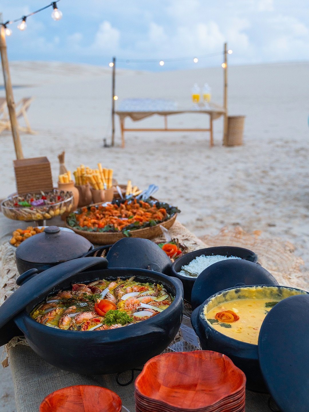 Mehrere traditionelle Tontöpfe mit Meeresfrüchten – darunter Garnelen-Moqueca und eine goldgelbe Cremesoße – auf einem rustikalen Tisch im weißen Sand der Dünen unter Lichterketten in der Abenddämmerung