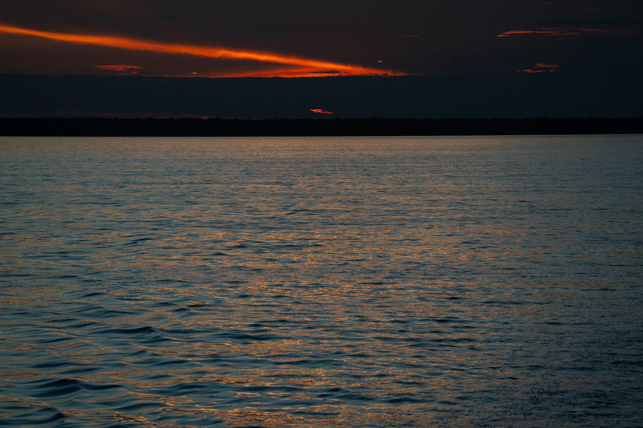 Deep red and orange streaks of cloud glow above a wide dark river at dusk on the Tucano Amazon river cruise, with a low forested horizon silhouetted beneath a near-black sky