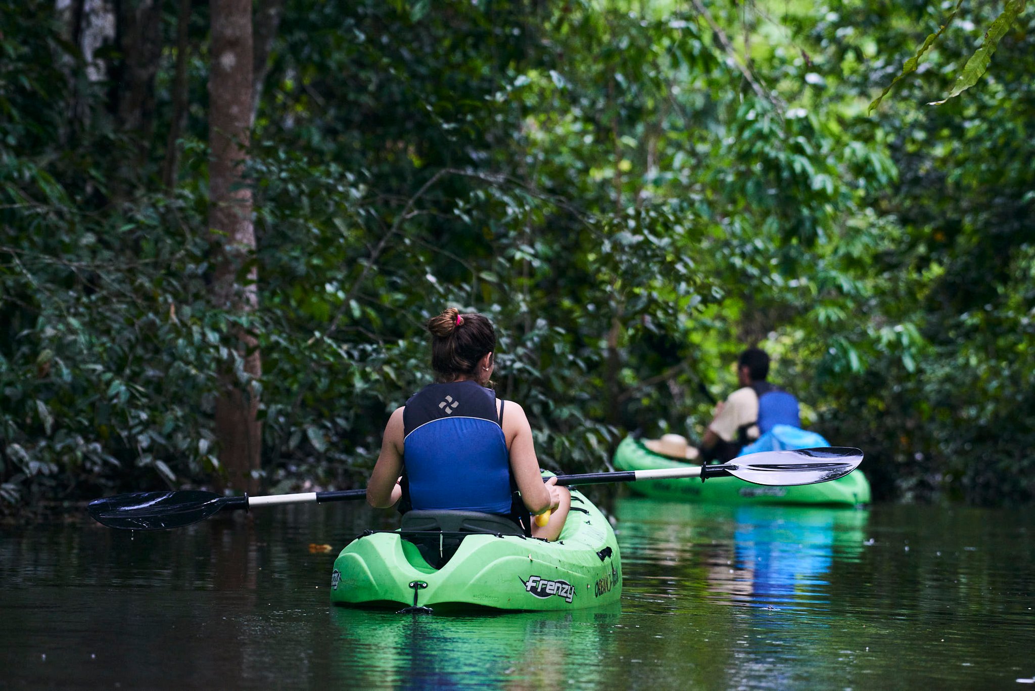 Two kayakers paddling green sit-on-top kayaks through a narrow, dark-watered channel lined with dense rainforest canopy on a tucano amazon river cruise igapó excursion