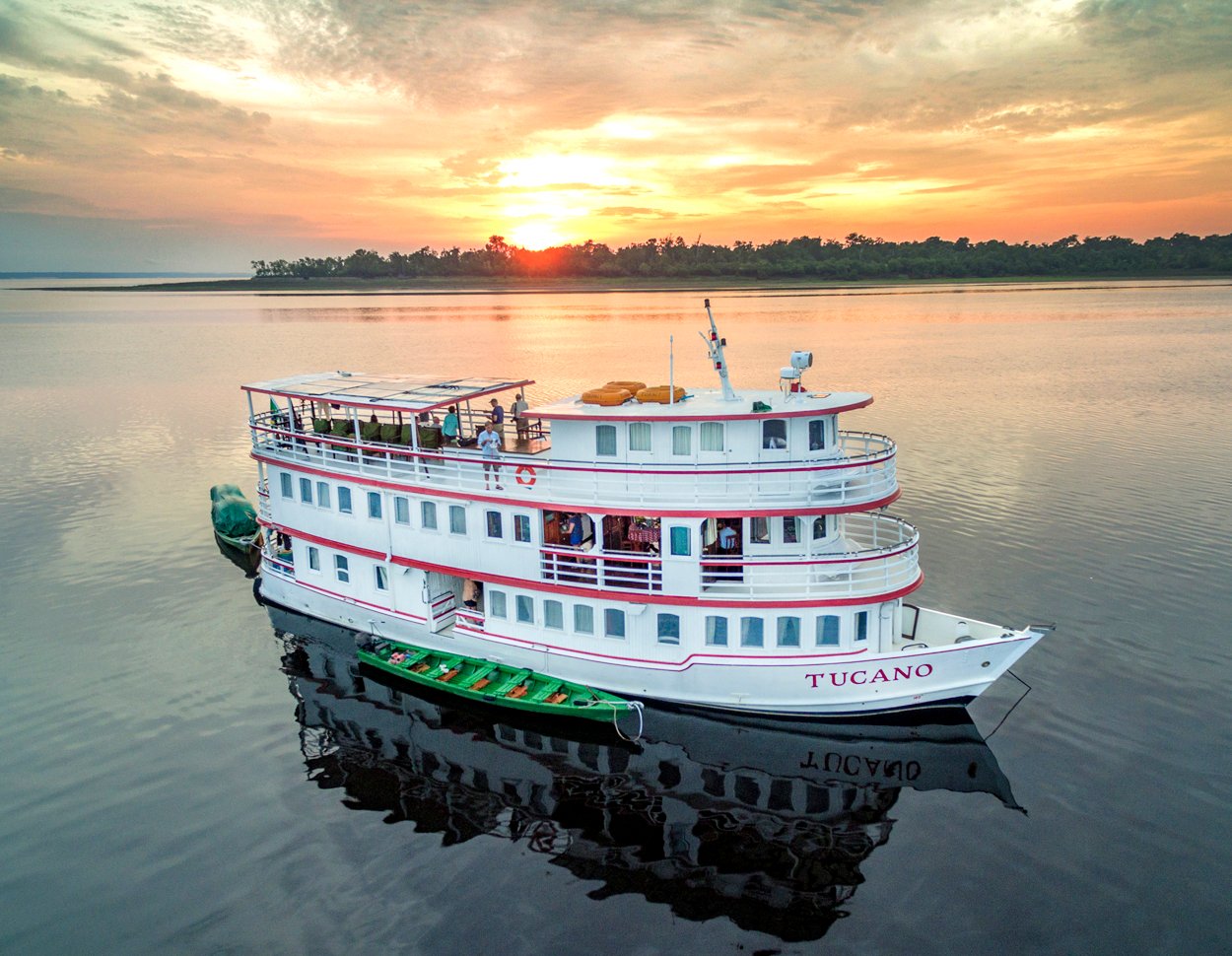 The M/Y Tucano, a white three-deck river cruise vessel, anchored on a glassy dark-water river at sunset, with a green wooden canoe alongside and passengers on the upper deck, forested shoreline silhouetted against an orange sky