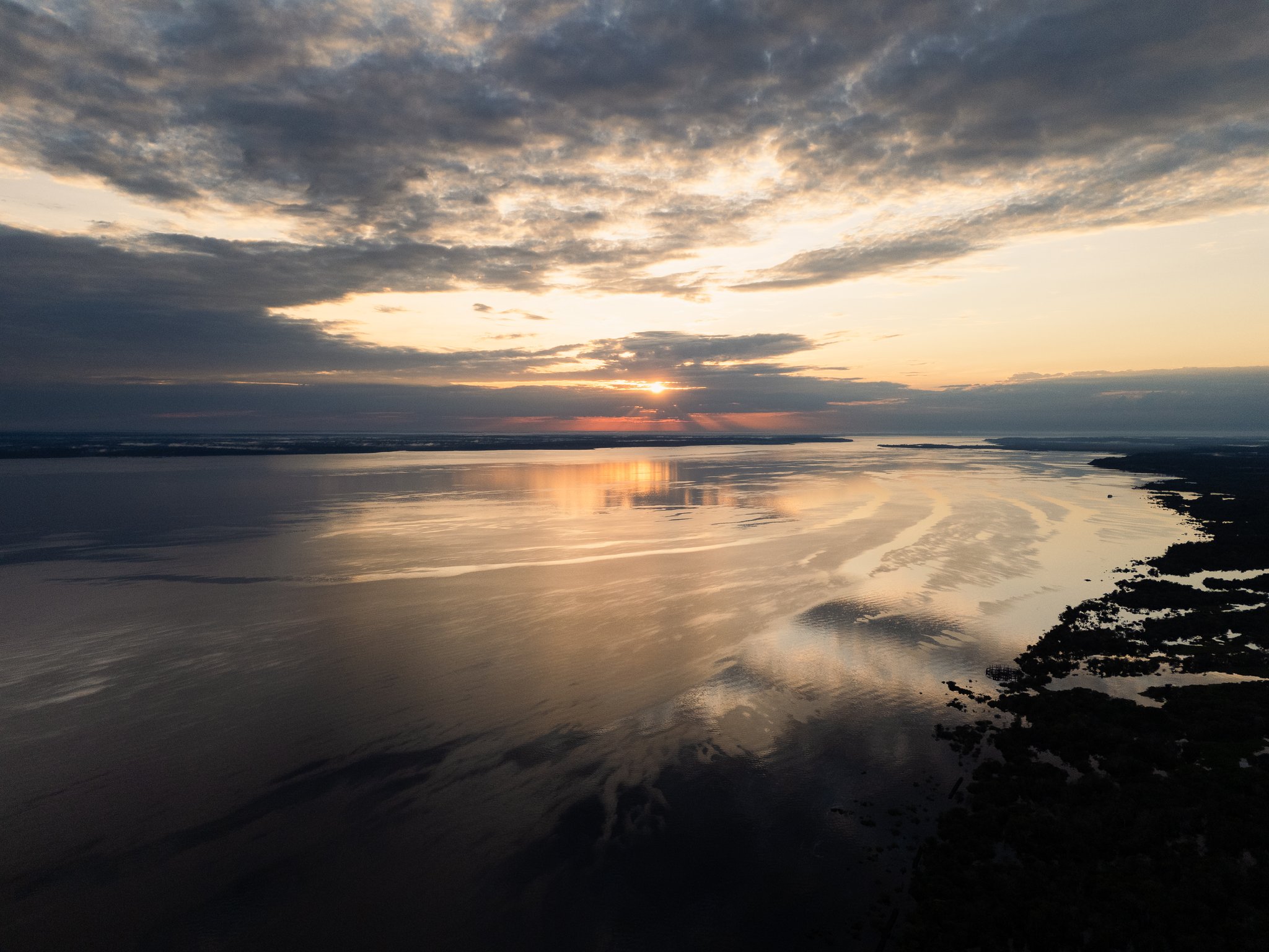 Aerial view of a vast Amazonian river at sunset on the tucano amazon river cruise, with golden light reflecting off the calm dark water and flooded vegetation silhouetted along the right bank beneath a dramatic cloud-filled sky
