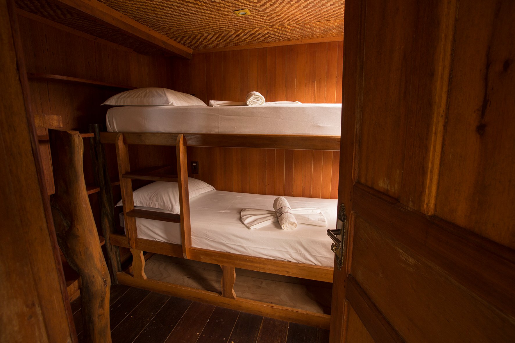 Small but considered, fresh linens and a wood-warm cabin after a long day on the river. Wooden bunk beds with crisp white linens and rolled towels inside a compact Amazon jungle cruise Brazil cabin with timber-panelled walls and woven ceiling