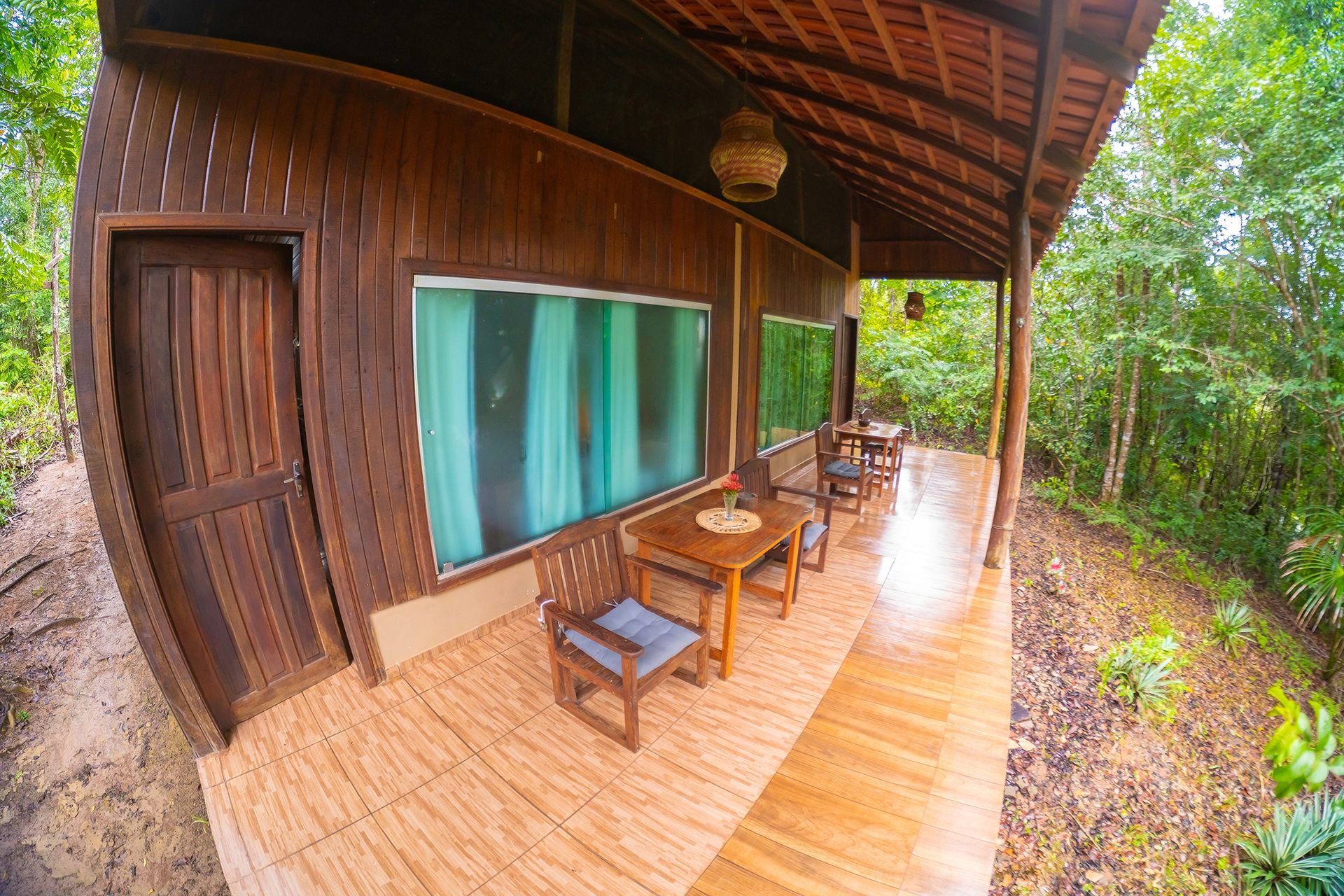 Het oerwoud begint precies waar de tegels ophouden. Een overdekte houten veranda van Amazon Turtle Lodge met houten stoelen, gekussende zitplaatsen en kleine eettafels langs een donkerhouten bungalow, omringd door dicht regenwoudgroen