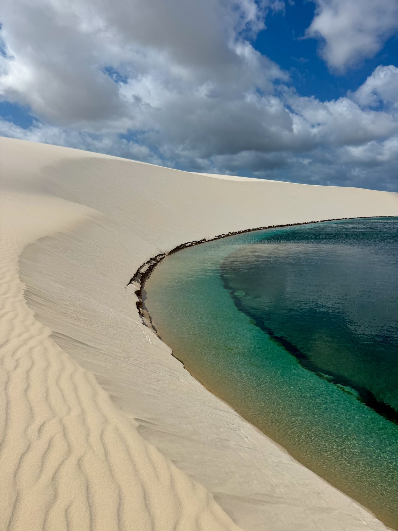 A crystal-clear turquoise lagoon nestled against a sweeping white sand dune under a dramatic cloudy sky, part of the Lençóis Maranhenses Full Itinerary experience