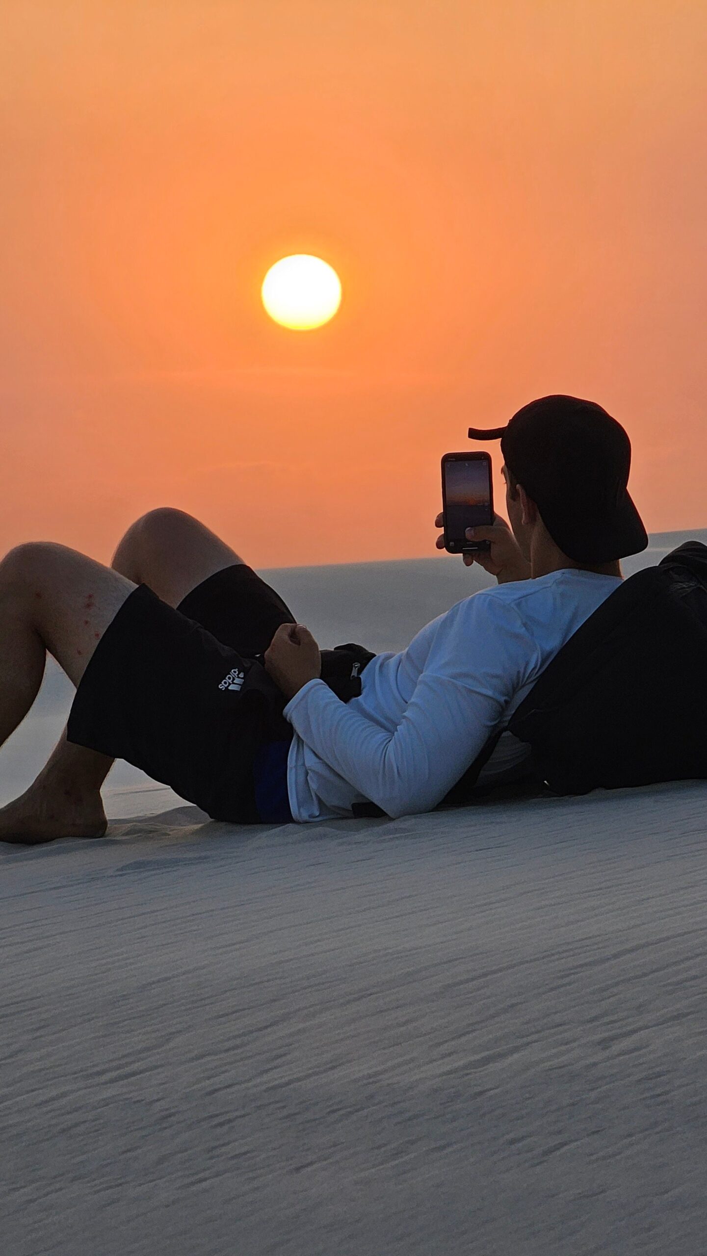 Ein Reisender liegt auf einer weißen Sanddüne in Lençóis Maranhenses und fotografiert einen großen orangefarbenen Sonnenuntergang mit seinem Smartphone