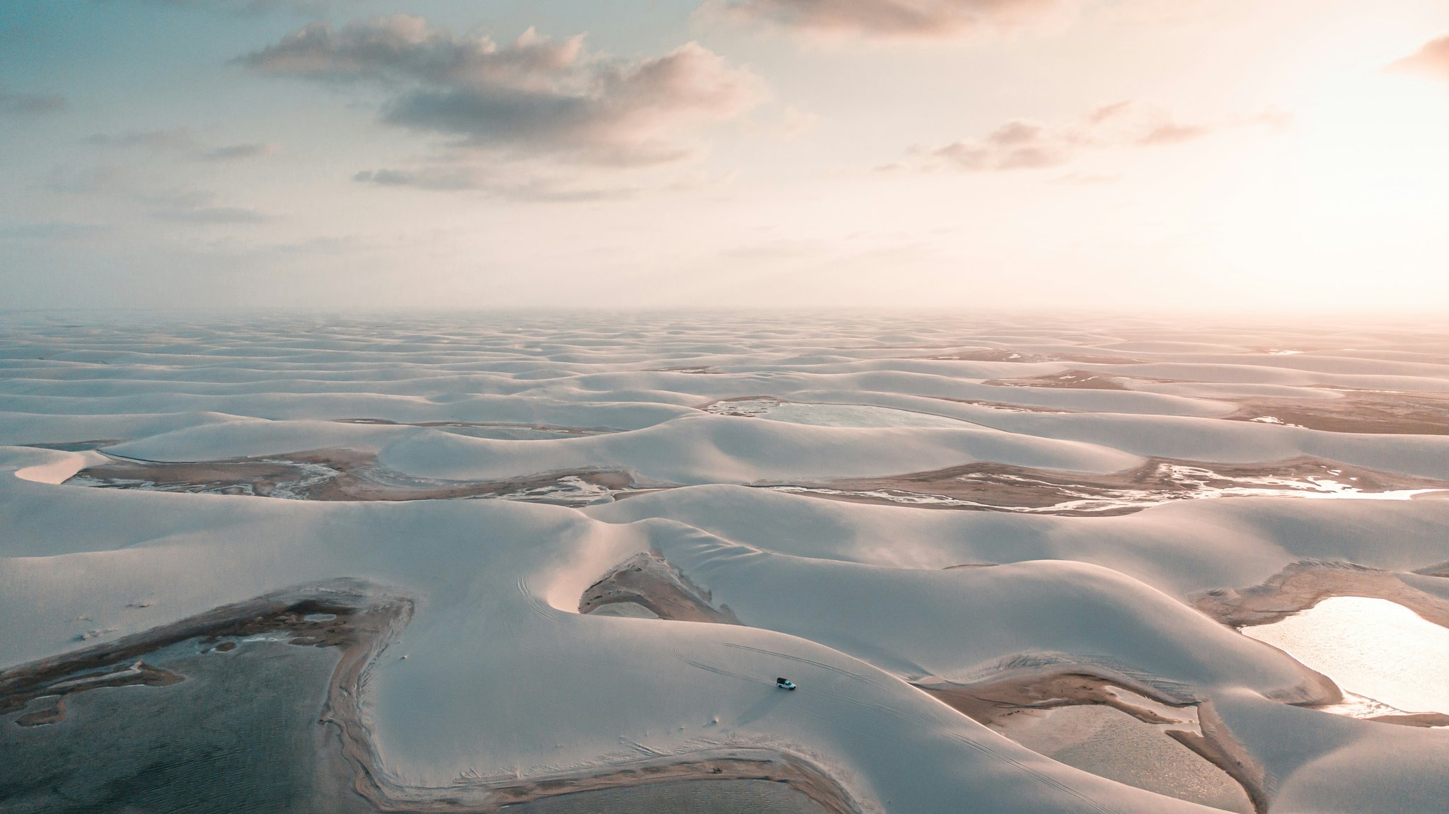 Weiße Dünen bis zum Horizont, ein paar Lagunen und ein winziges Fahrzeug, das alles in Relation setzt. Luftaufnahme des Lençóis Maranhenses Trek ab Atins mit einem einzelnen Geländewagen auf weißen Dünen, die von flachen Lagunen durchzogen sind, unter einem warm leuchtenden Himmel