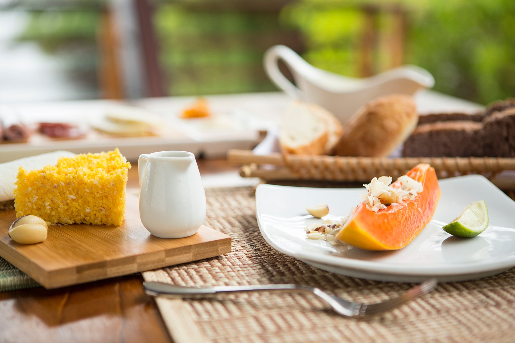 Rijpe papaja, zelfgebakken cake en de jungle als achtergrond, geen slecht begin van de dag. Een verse papayawig met noten en limoen naast een plak goudgeel maïsbrood op een bamboeplank, geserveerd als ontbijt bij Mirante do Gavião, met een broodmand op de achtergrond