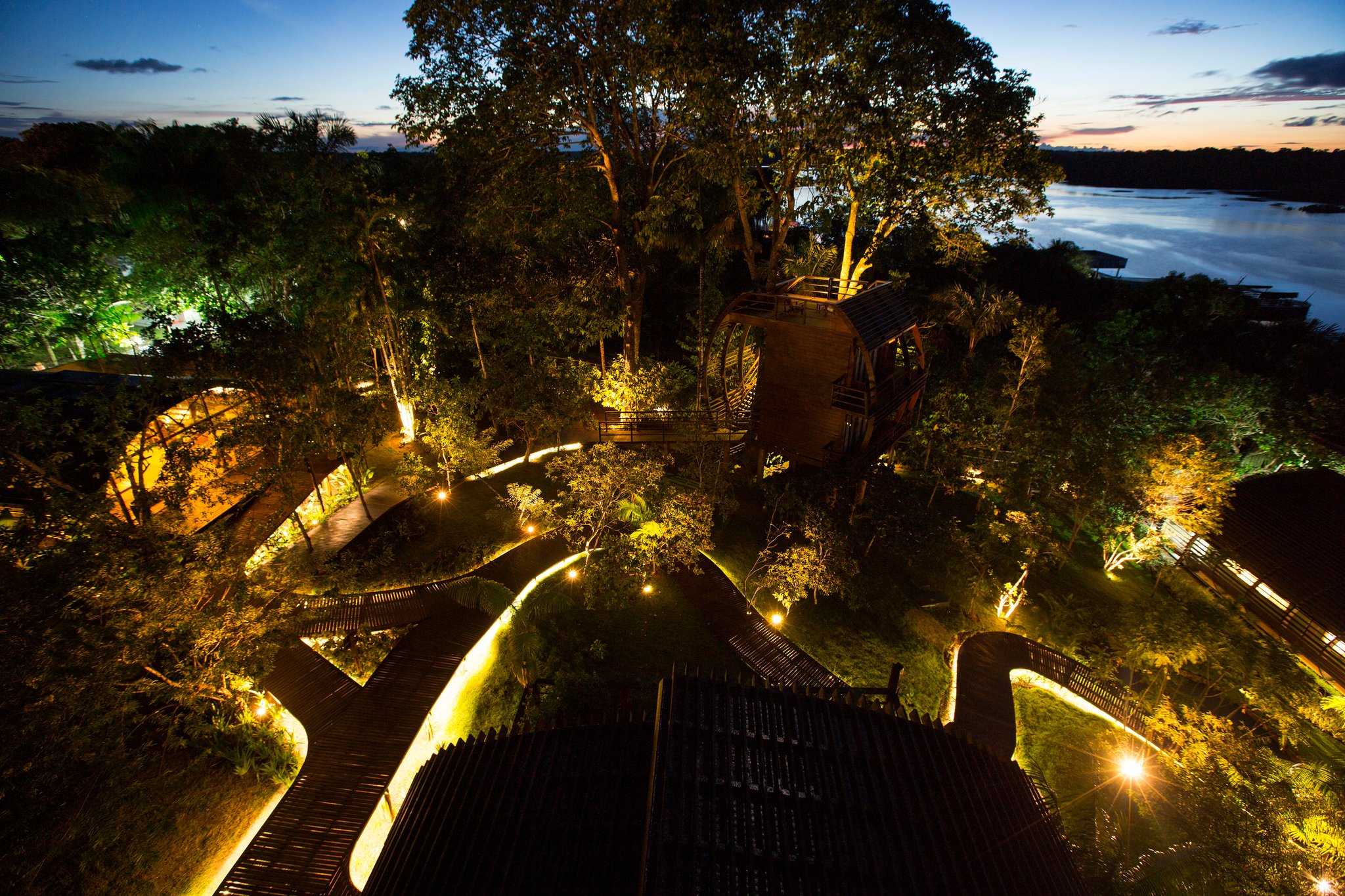 Luchtfoto van Mirante do Gavião amazon lodge bij schemering, met verlichte houten boardwalks die door het regenwoudkronendak slingeren, een cilindrisch boomhuis en de glinsterende Rio Negro op de achtergrond