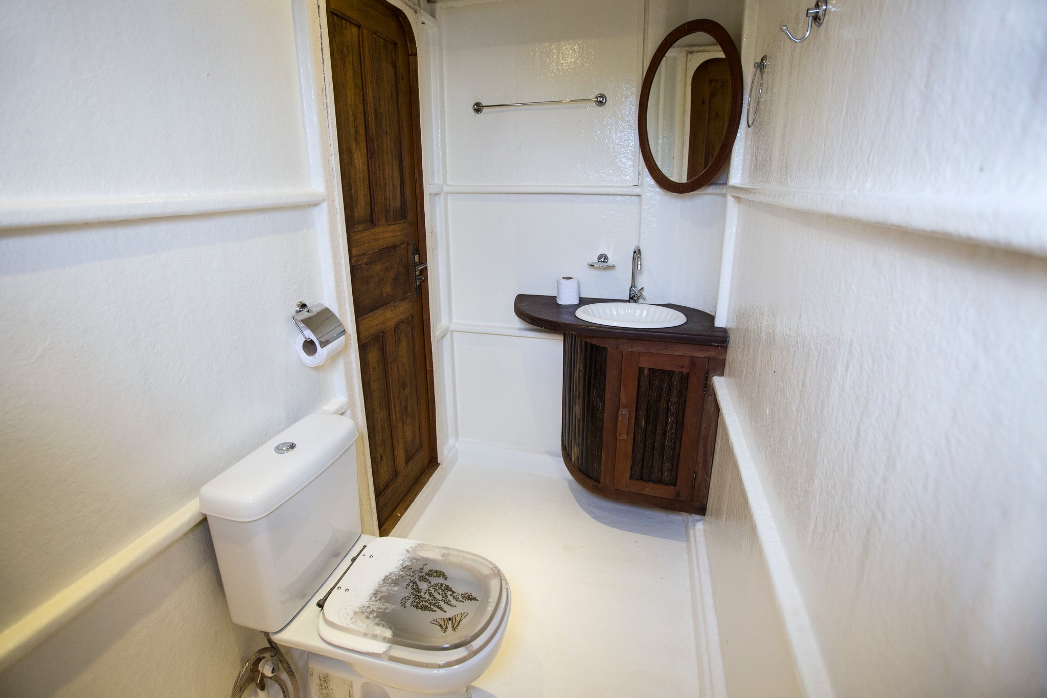 Small but thoughtfully fitted out, right down to the wooden details. Compact private en-suite bathroom aboard an Amazon jungle cruise Brazil vessel, featuring a curved wooden vanity unit, round mirror, and decorated toilet seat in a white-painted cabin