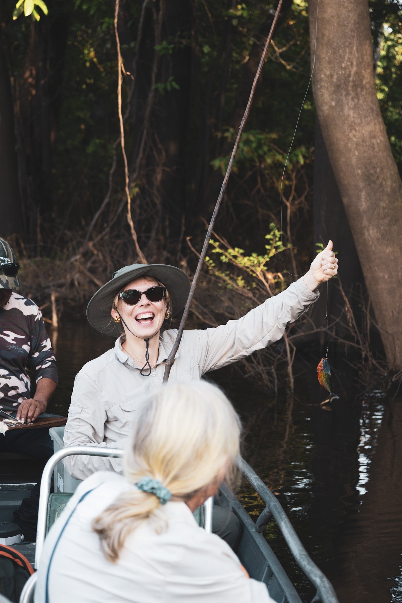 Eerste vangst, grootste grijns. Piranhaën doen dat met je. Een lachende vrouw met zonnehoed houdt een net gevangen visje omhoog vanuit een gemotoriseerde kano tijdens een piranhavisexcursie bij de Amazon Turtle Lodge op een donker boskanaal