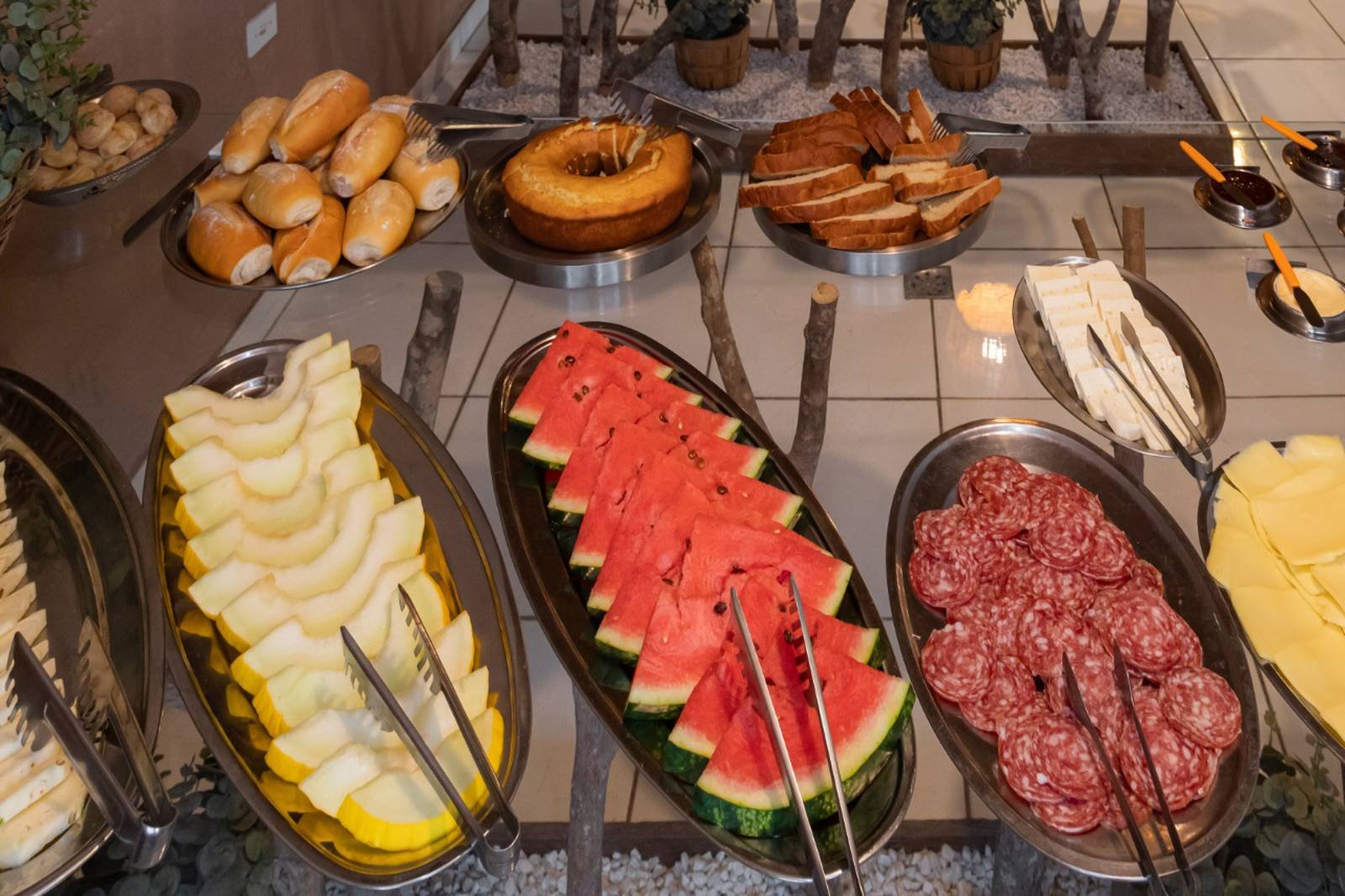 Een uitgebreid ontbijtbuffet op een budget Pantanal tour, met schijven watermeloen, honingmeloen, salamiplakjes, kaas, broodjes, een ringcake, geroosterd brood en dipsauzen in roestvrijstalen schalen