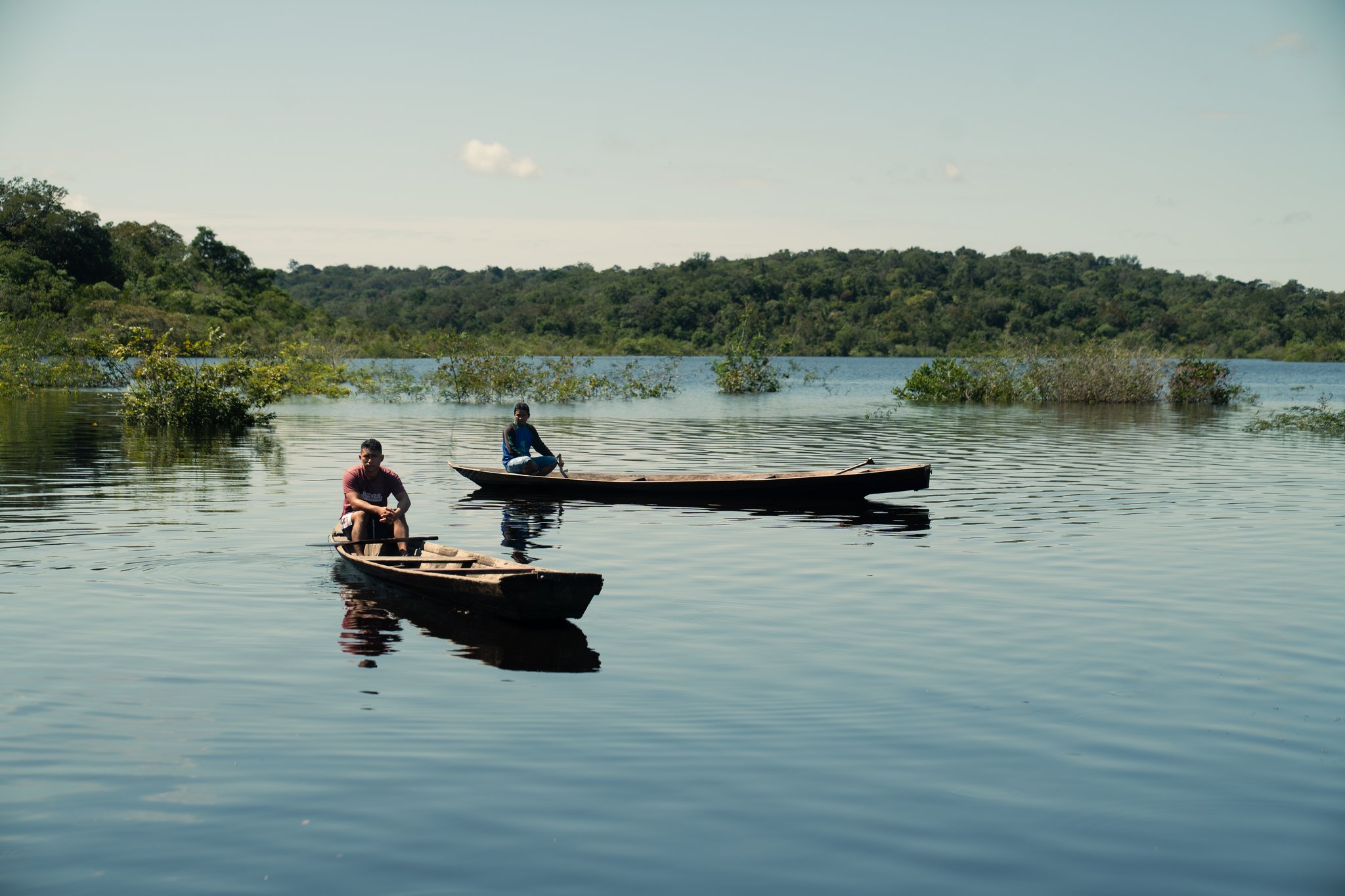 Two local guides rest in traditional wooden dugout canoes on a calm river surrounded by flooded vegetation and dense rainforest, an unhurried moment that captures the spirit of community based tourism on Amazonian waterways