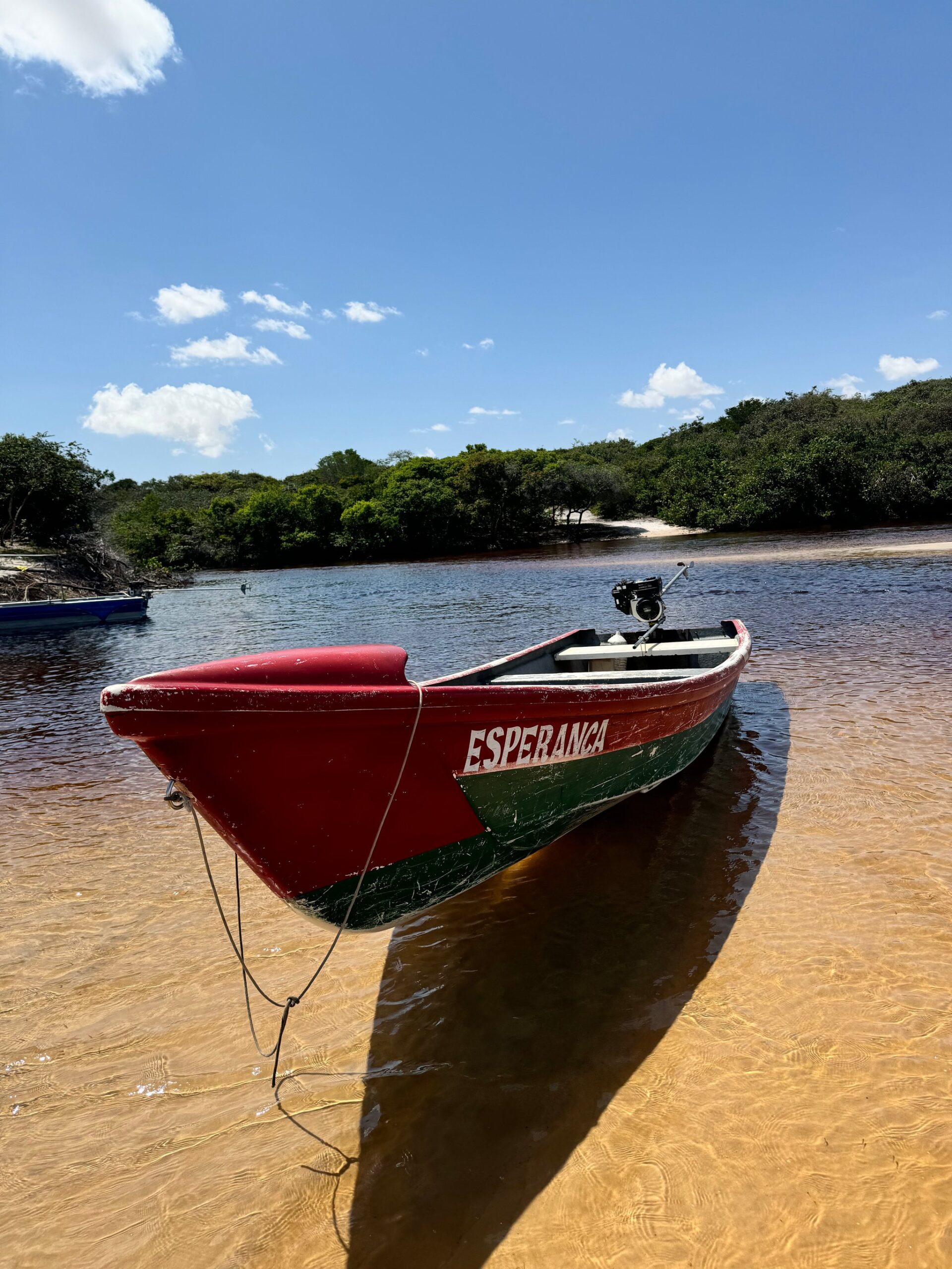 A red and green wooden boat named Esperança moored at a sandy riverbank, with mangrove-fringed water and blue sky behind — part of the Lençóis Maranhenses full itinerary river experience