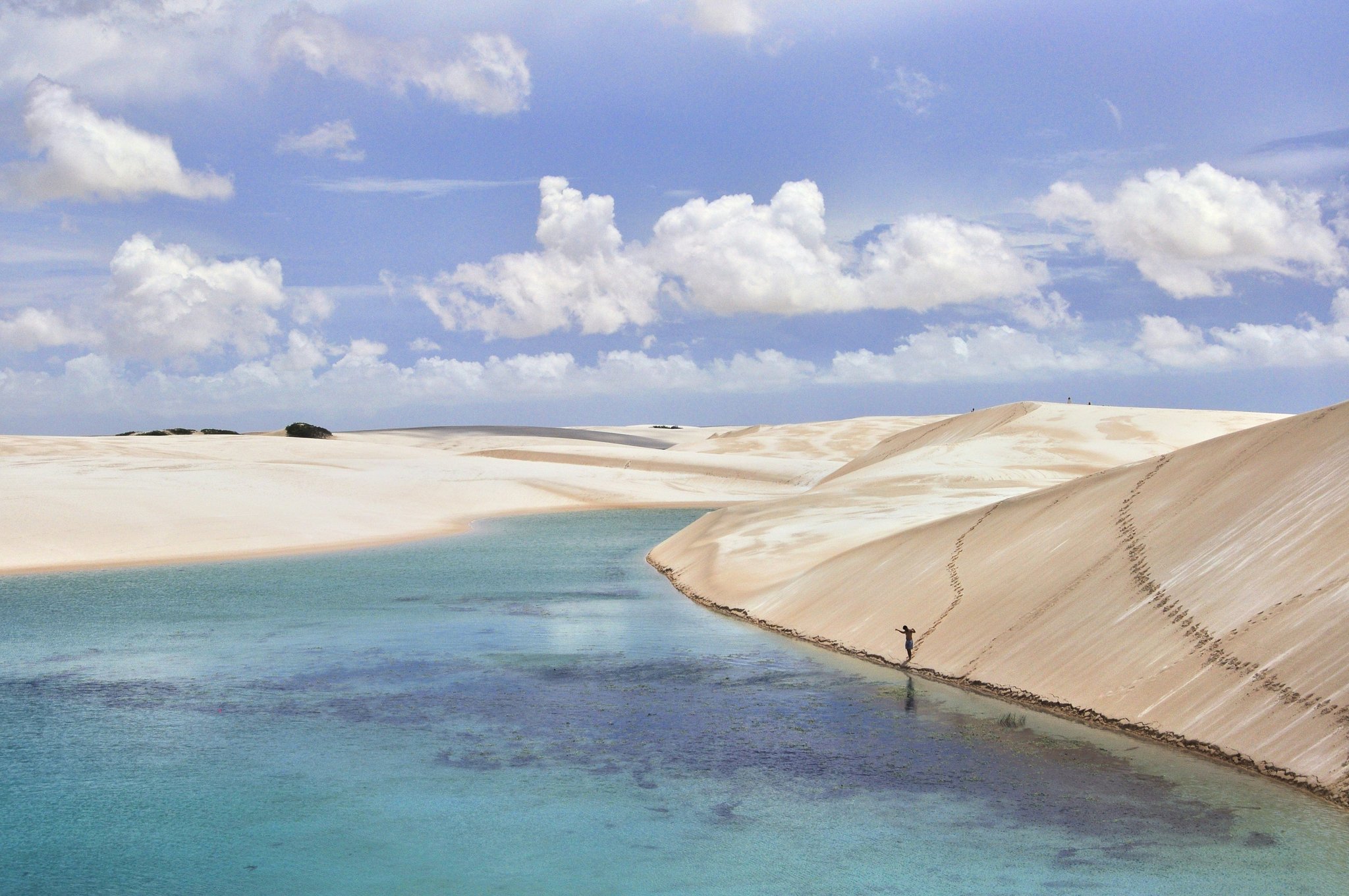 A lone figure wades along the base of a sweeping white sand dune beside a turquoise lagoon in Lençóis Maranhenses Full Itinerary country, under a wide blue sky with cumulus clouds