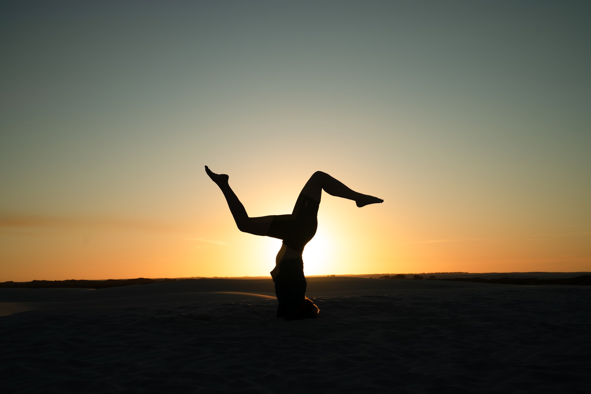 Silhouette of a person performing a headstand with legs split wide open on the sand dunes at golden-hour sunset, part of a Lençóis Maranhenses full itinerary experience