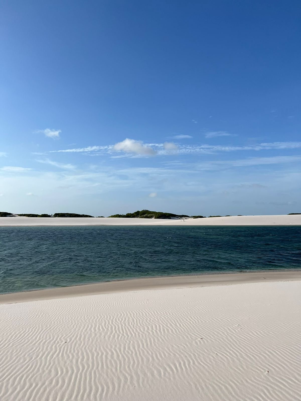 Breite Süßwasserlagune, flankiert von gewellten weißen Sanddünen unter klarem blauem Himmel in Lençóis Maranhenses Santo Amaro, mit vereinzelter grüner Vegetation am fernen Dünenkamm