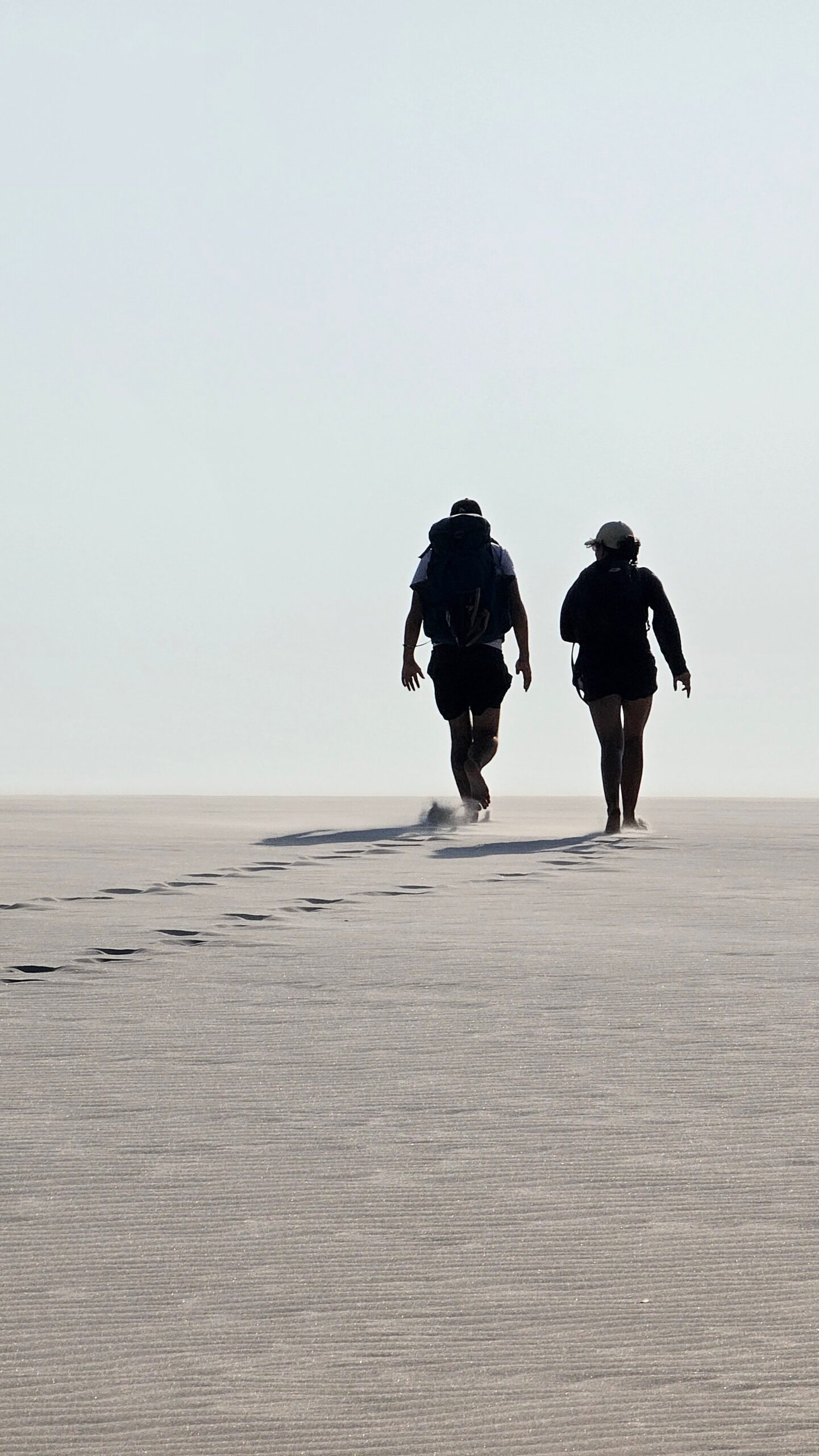 Weißer Sand bis zum Horizont, und ihr seid die Einzigen, die ihn durchqueren. Zwei Wanderer laufen auf dem Lençóis-Maranhenses-Trek von Atins über eine weite, flache Weißsandfläche, ihre Fußspuren verlieren sich im dunstigen Horizont
